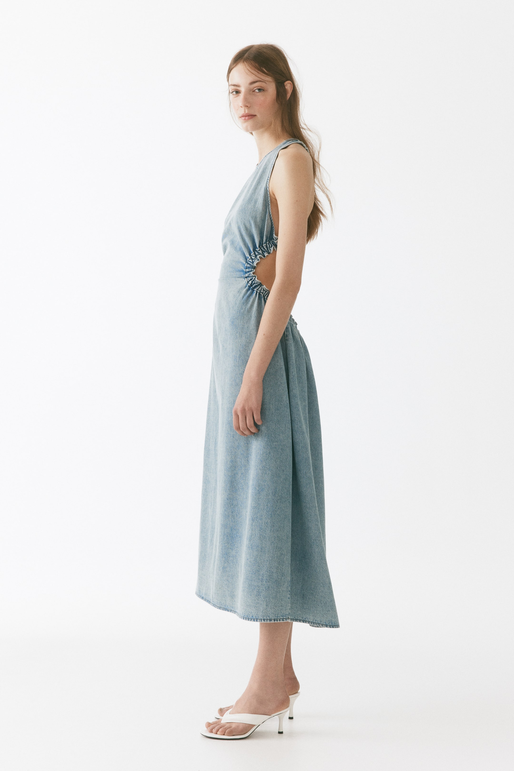 View larger image: Presenting a faded blue denim sleeveless midi dress from a side angle, this piece showcases circular side cut-outs with gathered ruching details at the waist, seamlessly flowing into an A-line skirt.
