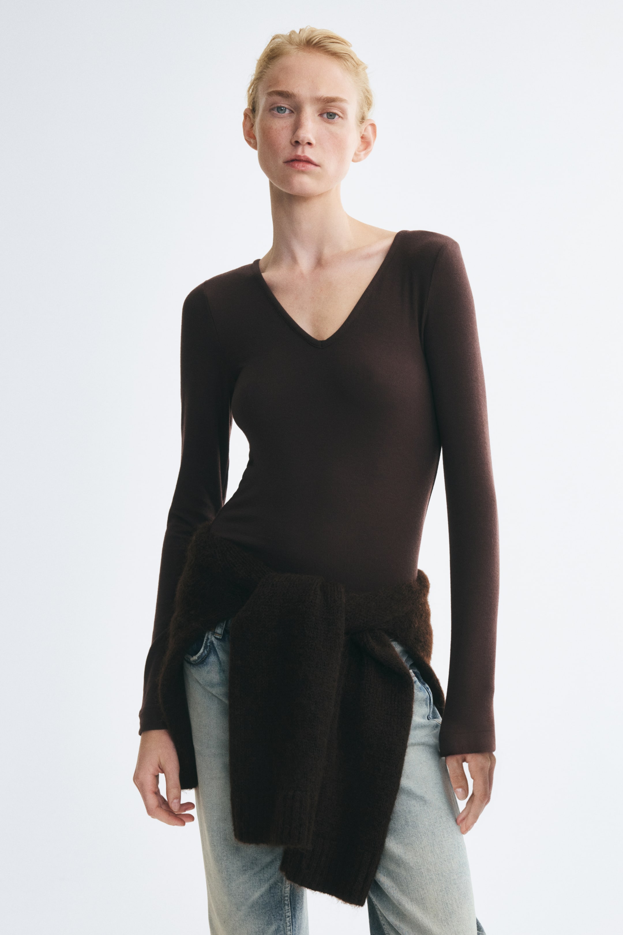 View larger image: A dark brown fitted long-sleeve top with a V-neck is worn by the model. The top is styled with light-wash blue jeans and a matching dark brown knit sweater tied around the waist.