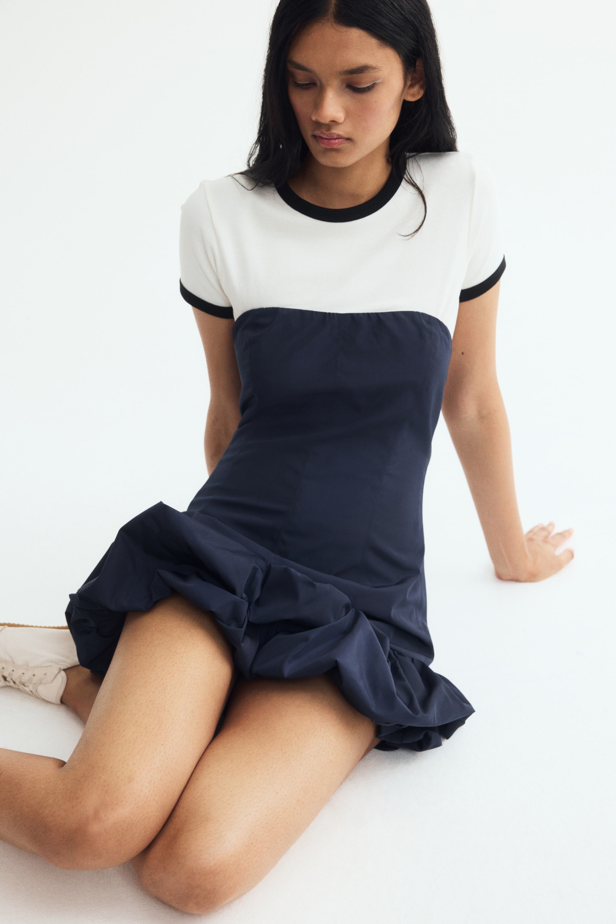 View larger image: A two-tone mini dress is being worn by the model while seated. The dress features a white short-sleeved top with black ringer trim at the crew neck and sleeves, joined to a dark navy blue fitted body with a voluminous bubble hem.