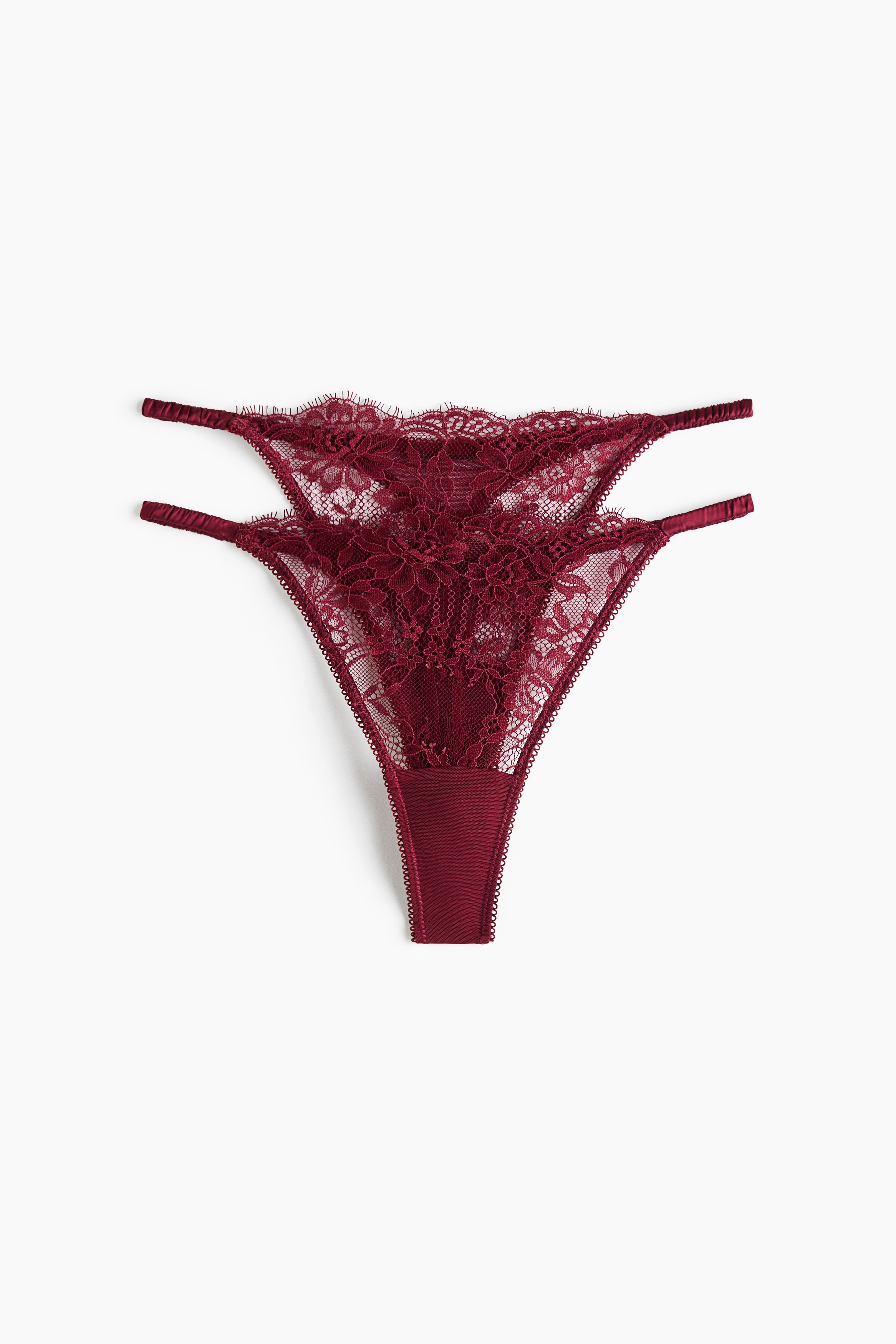 La modelo lleva un tanga de encaje y satén en rojo oscuro, con cintura de tiro bajo, finas tiras laterales y ribete de encaje con acabado de pestañas. También lleva un bralette a juego de encaje en rojo oscuro, parcialmente visible en la parte superior de la imagen.