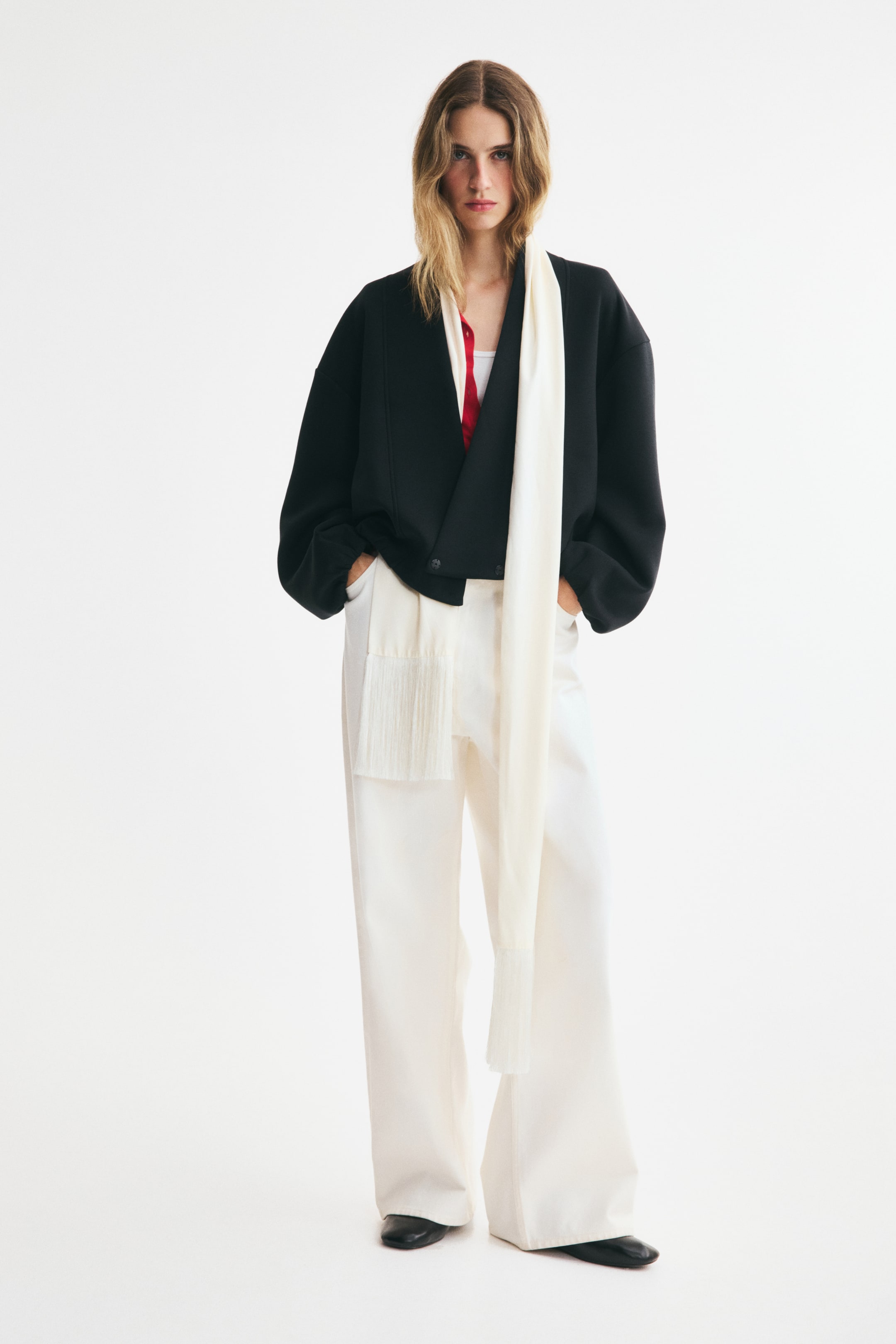 View larger image: This front-facing black wrap jacket is showcasing a relaxed yet structured fit with a V-neckline and voluminous sleeves. It is being worn layered over a red shirt and styled with white wide-leg trousers and a long, cream-coloured scarf featuring fringed edges.