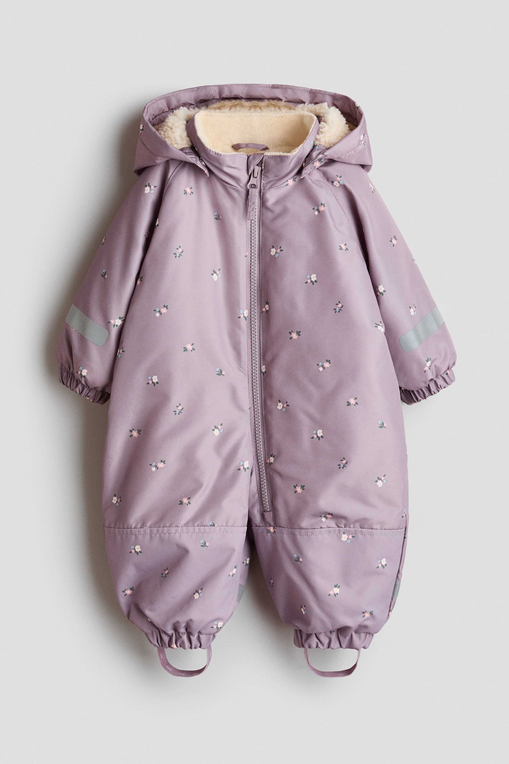 Agrandir l'image: Une combinaison matelassée tout-en-un est présentée de face, dans une couleur lilas avec un petit imprimé floral. Elle est dotée d’une capuche amovible doublée en fausse fourrure de mouton et d’un col montant, d’une fermeture zippée intégrale sur le devant, de bandes réfléchissantes sur les manches, de genoux renforcés, de poignets et d’ourlets élastiqués, ainsi que de sous-pieds amovibles.