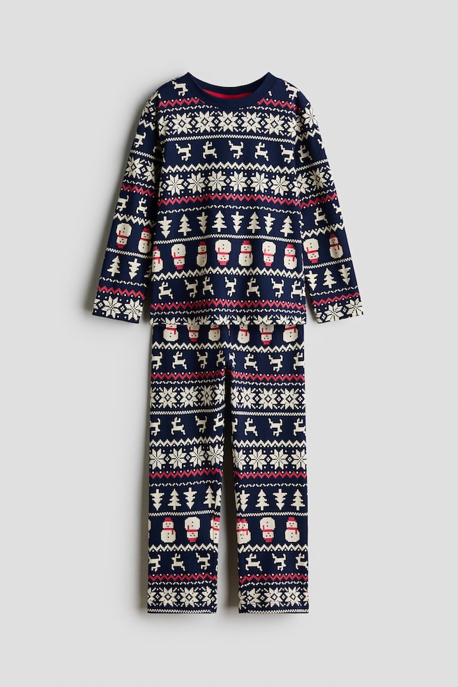A front-facing two-piece pyjama set features a matching long-sleeve top with a round neckline and straight-leg trousers in navy blue cotton jersey. Both pieces feature an all-over knit-style pattern of snowflakes, reindeer, pine trees, and snowmen in white and red. - 1