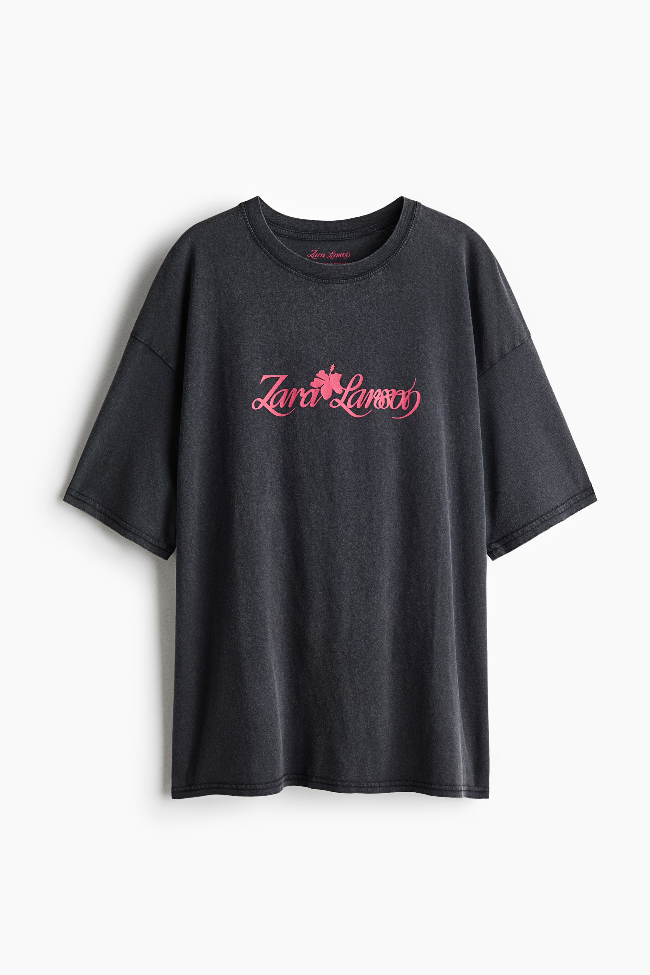 Grotere afbeelding bekijken: Een oversized donkergrijs katoenen T-shirt hangt met de voorkant naar voren en heeft een geribde ronde hals, afhangende schouders en brede korte mouwen. Op de borst staat een roze print met de cursieve tekst ‘Zara Larsson’ en een hibiscusbloem in het midden.