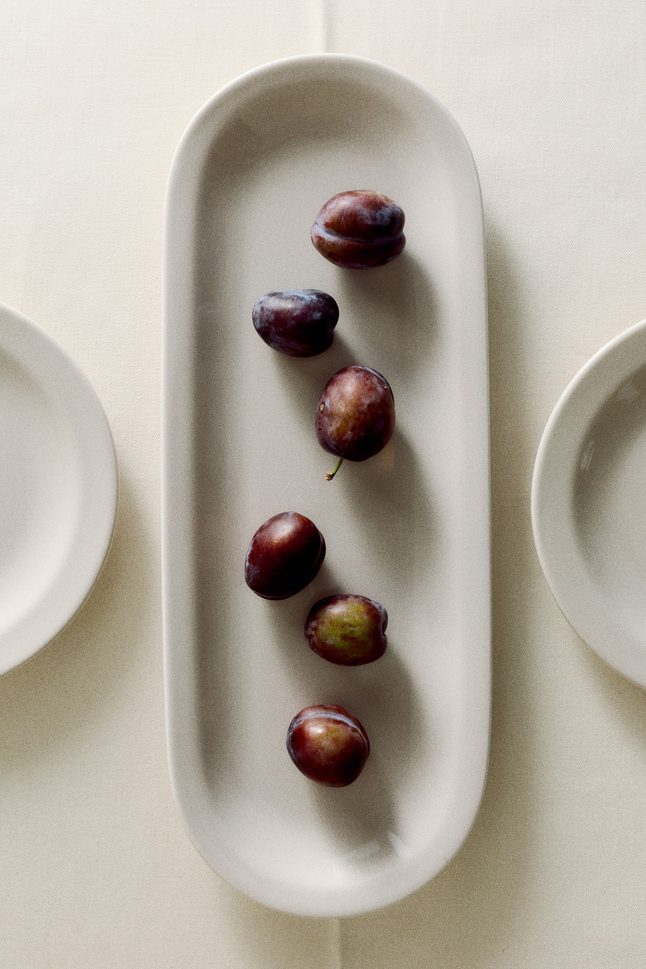 View larger image: An oval porcelain serving platter with a minimalist design and a narrow rim is shown from above, displaying six whole plums arranged in a line down its center. The off-white platter features a smooth matte finish and gently raised edges.