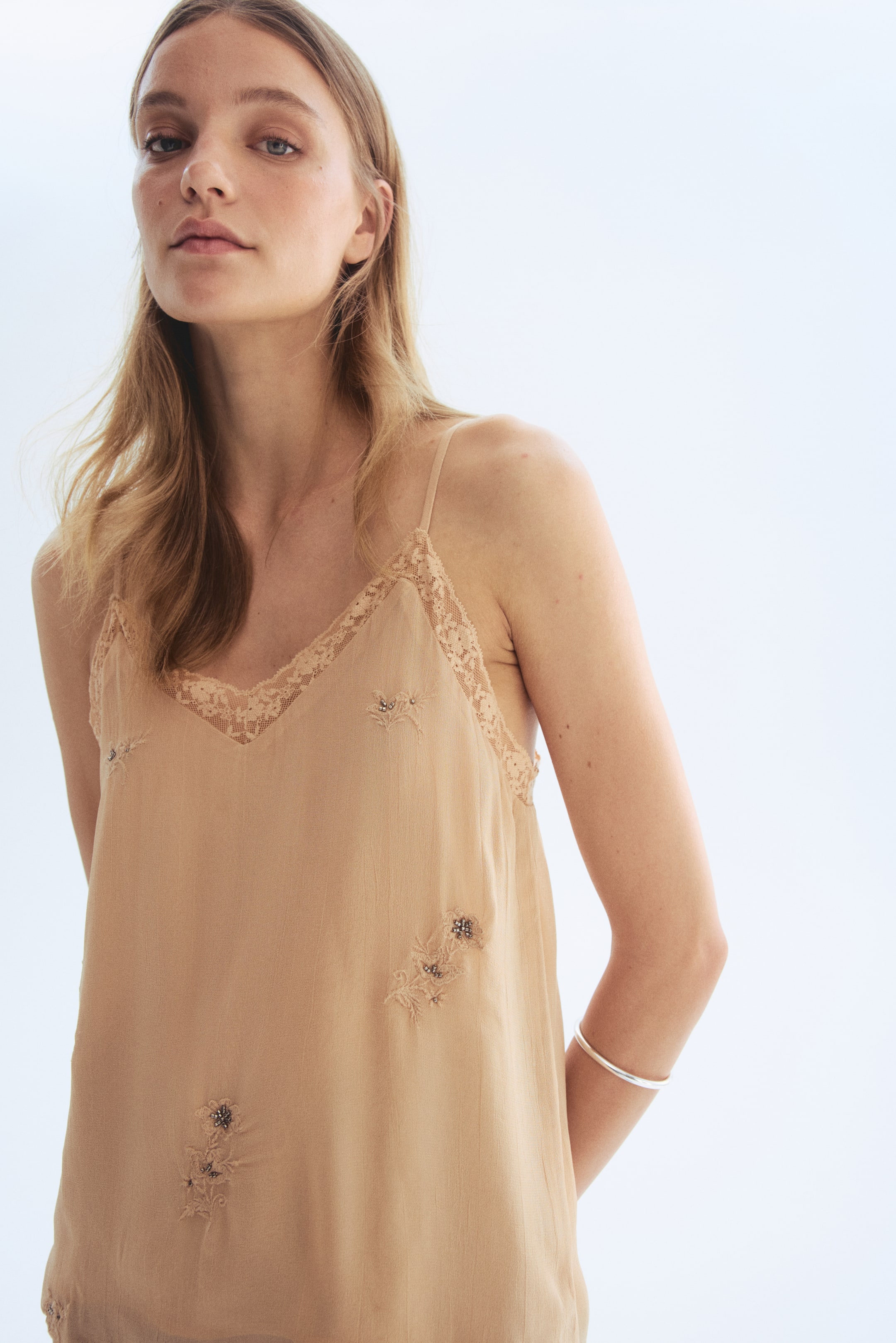 View larger image: A beige camisole top, facing forward, features a V-neckline, thin straps, and lace trim along the neckline and armholes, while embroidered floral patterns with small beads adorn its front.