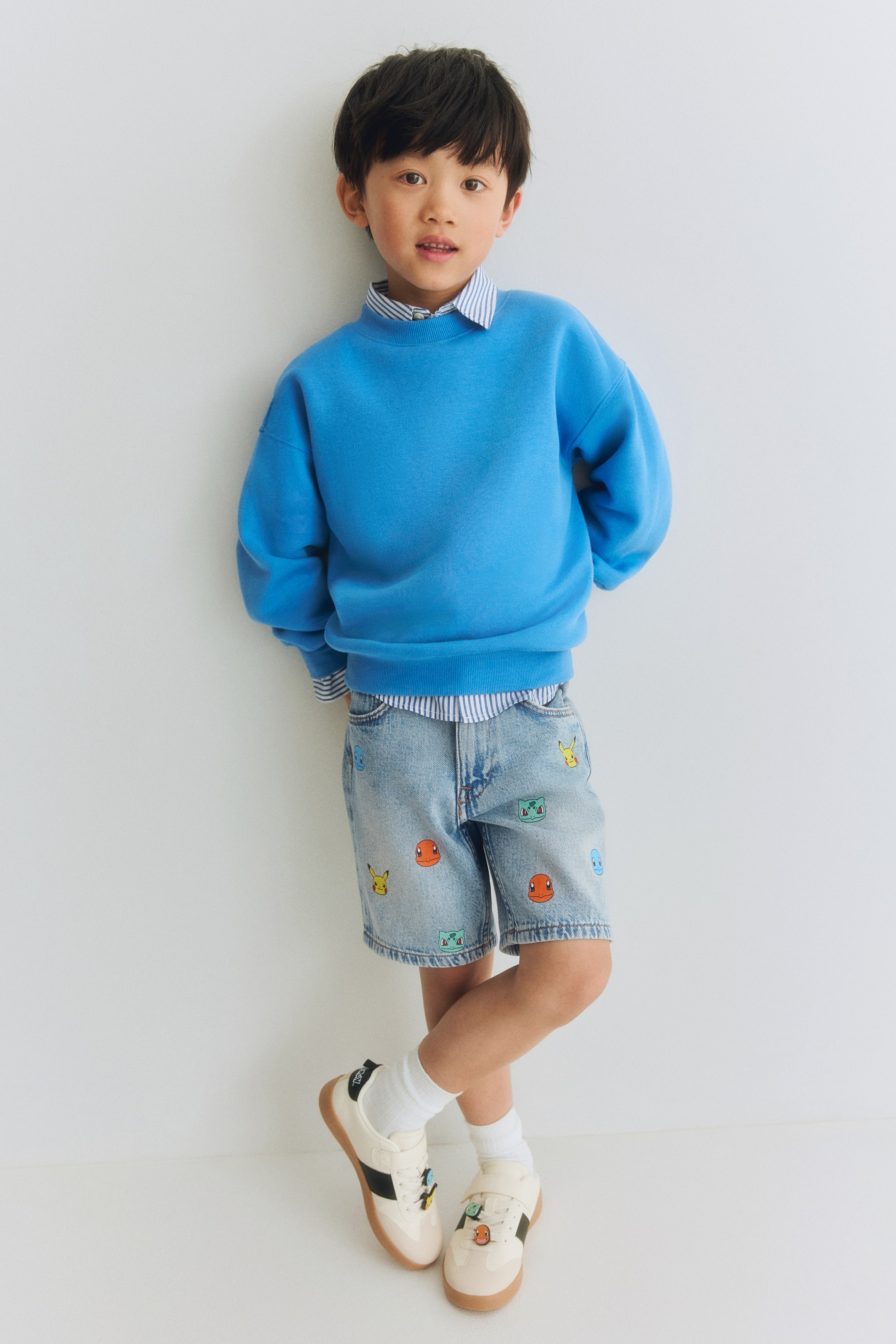 Grösseres Bild ansehen: Ein blaues Sweatshirt mit Rundhalsausschnitt wird getragen, das durch eine entspannte Passform, lange Ärmel mit gerippten Bündchen und einen gerippten Saum besticht, über einem gestreiften Hemd mit Kragen geschichtet.