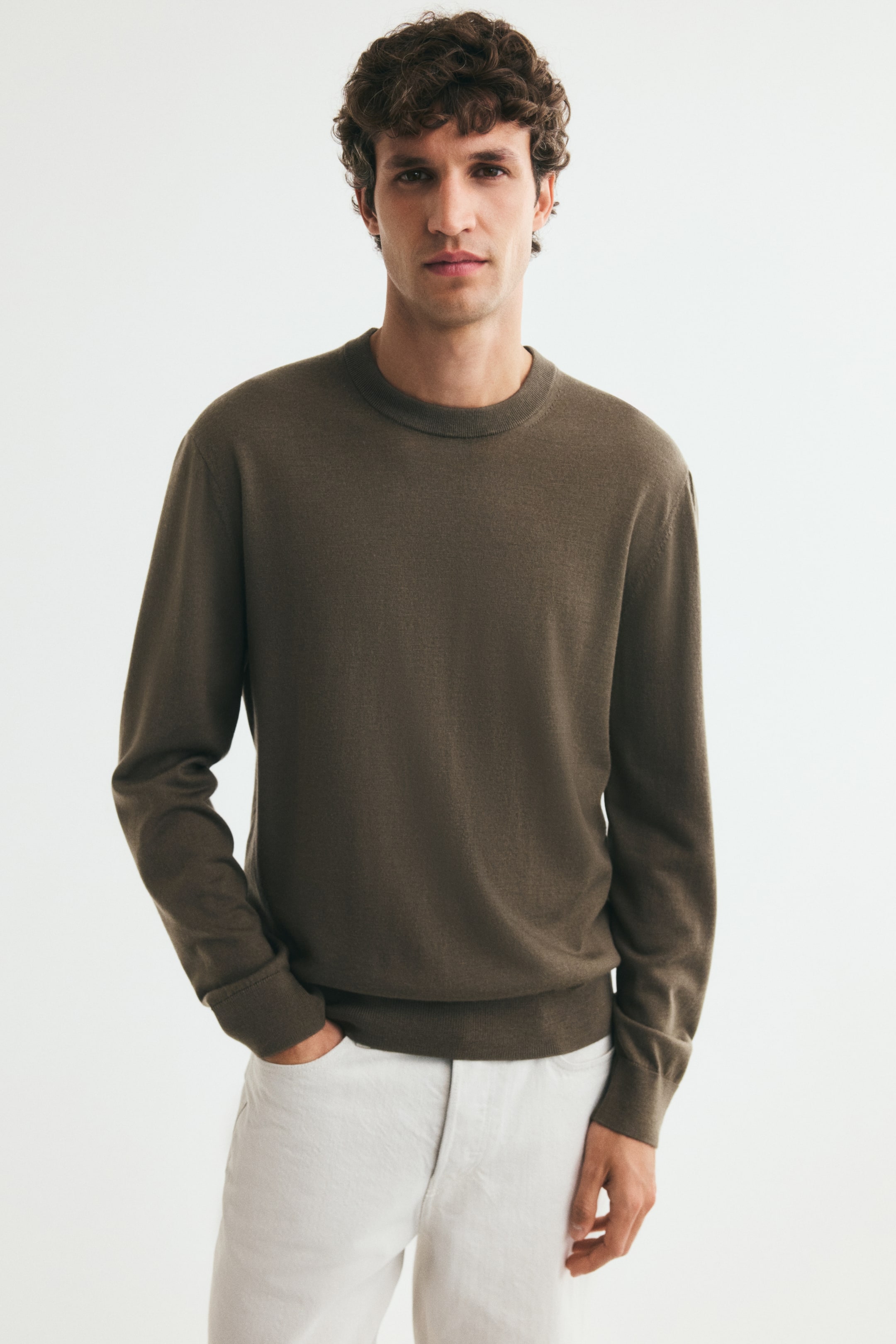 View larger image: A fine-knit, olive-brown jumper featuring a round, rib-trimmed neckline, long sleeves with ribbed cuffs, and a ribbed hem is being worn by the model. The classic-fit jumper is paired with light-coloured trousers, with the model resting one hand in a front pocket.