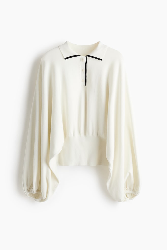 SuÃ©ter tipo polo tejido en color blanco roto, mostrado de frente, con cuello clÃ¡sico y cartera de tres botones detallada con un fino ribete negro en contraste. El diseÃ±o presenta mangas globo voluminosas y drapeadas con puÃ±os fruncidos y una pretina ancha de canalÃ© ajustada. - 1