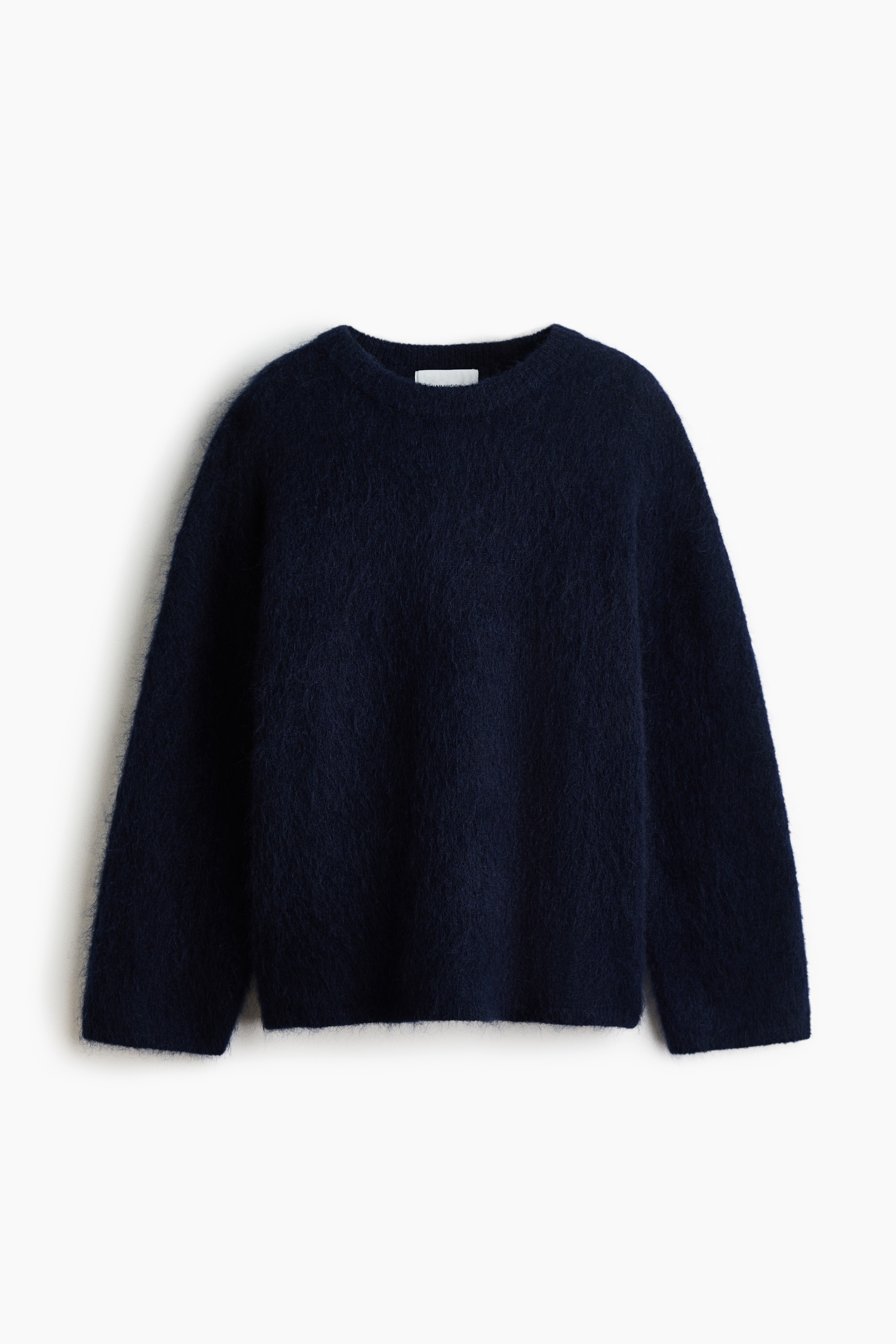 La modelo lleva un jersey de punto azul oscuro y afelpado, combinado sobre una camisa blanca con cuello y con vaqueros azules. El jersey de corte oversize presenta hombros caÃ­dos, cuello redondo de canalÃ© y puÃ±os acanalados.