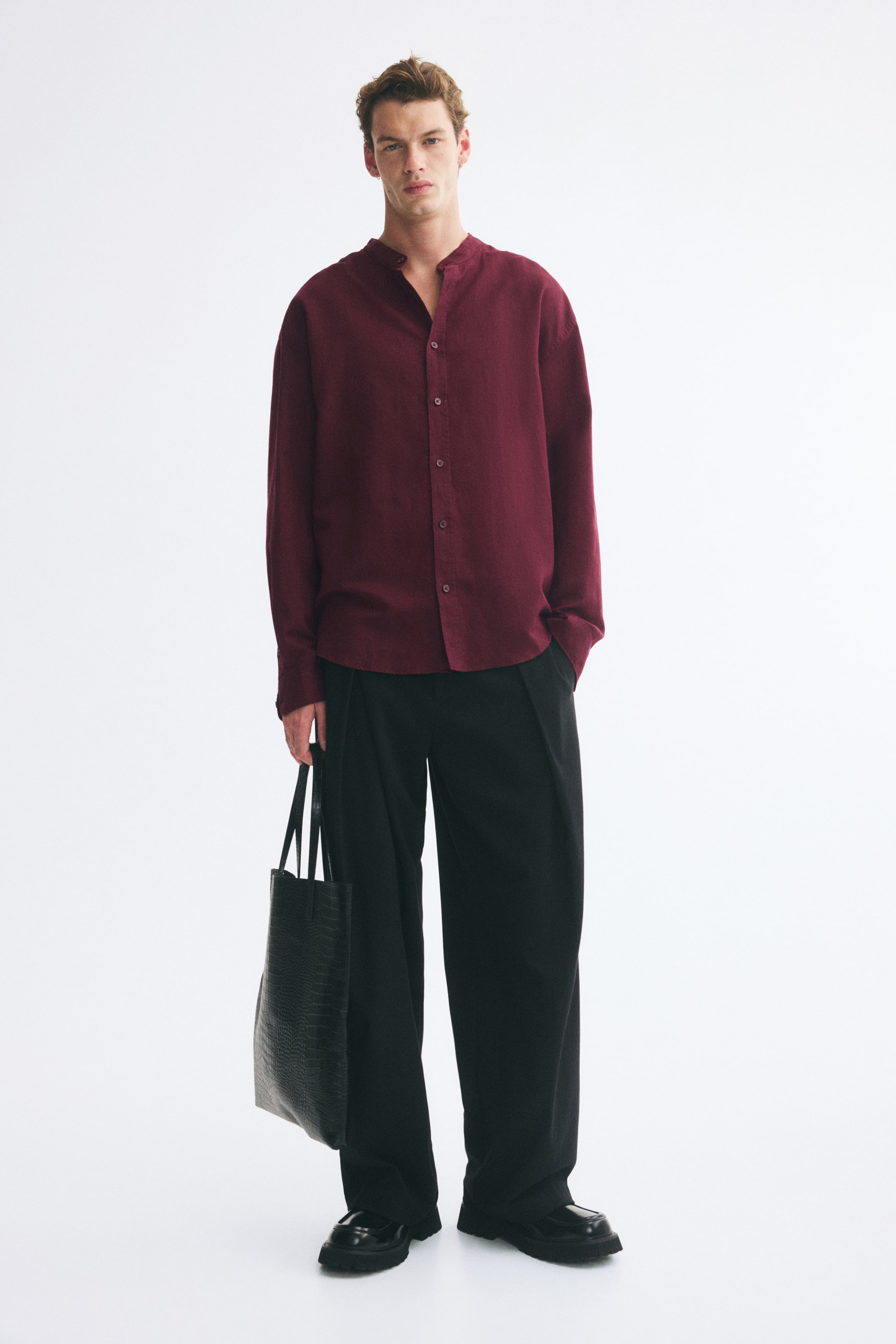 View larger image: A burgundy linen-blend shirt featuring a band collar, button-up front, and long sleeves is worn by the model. The relaxed-fit shirt is styled untucked over wide-leg black trousers, while the model is holding a black textured tote bag.