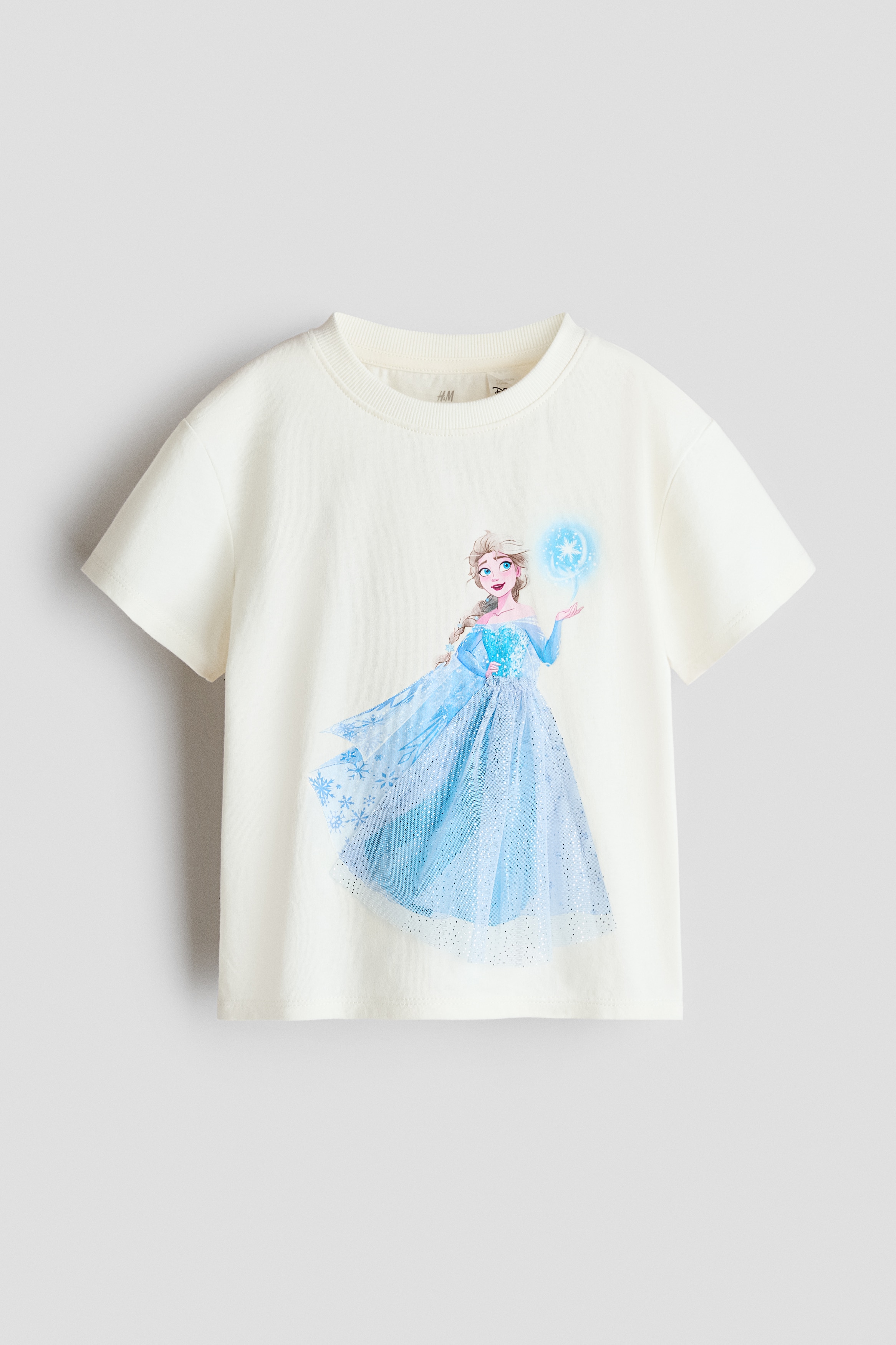 A cream-coloured, short-sleeved T-shirt featuring a graphic motif of a character in a blue gown is being worn by the model. The product is paired with light-wash, wide-leg denim jeans.