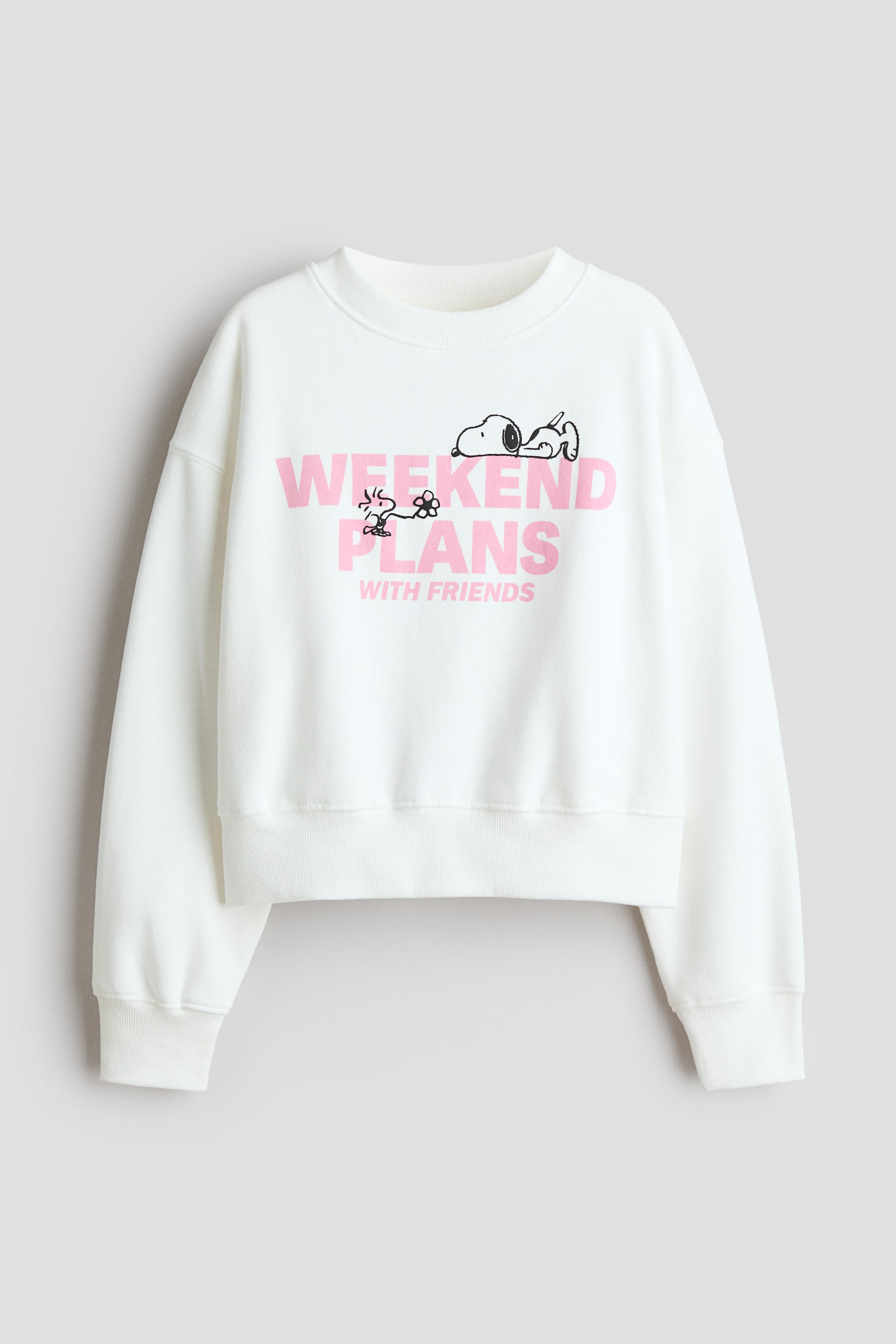 View larger image: A white crew neck sweatshirt featuring long sleeves with ribbed cuffs and a ribbed hem. The front displays a graphic print with the words "Weekend Plans" in pink, "With Friends" in smaller pink text, and a black outline illustration of Snoopy lying down with Woodstock holding a flower.