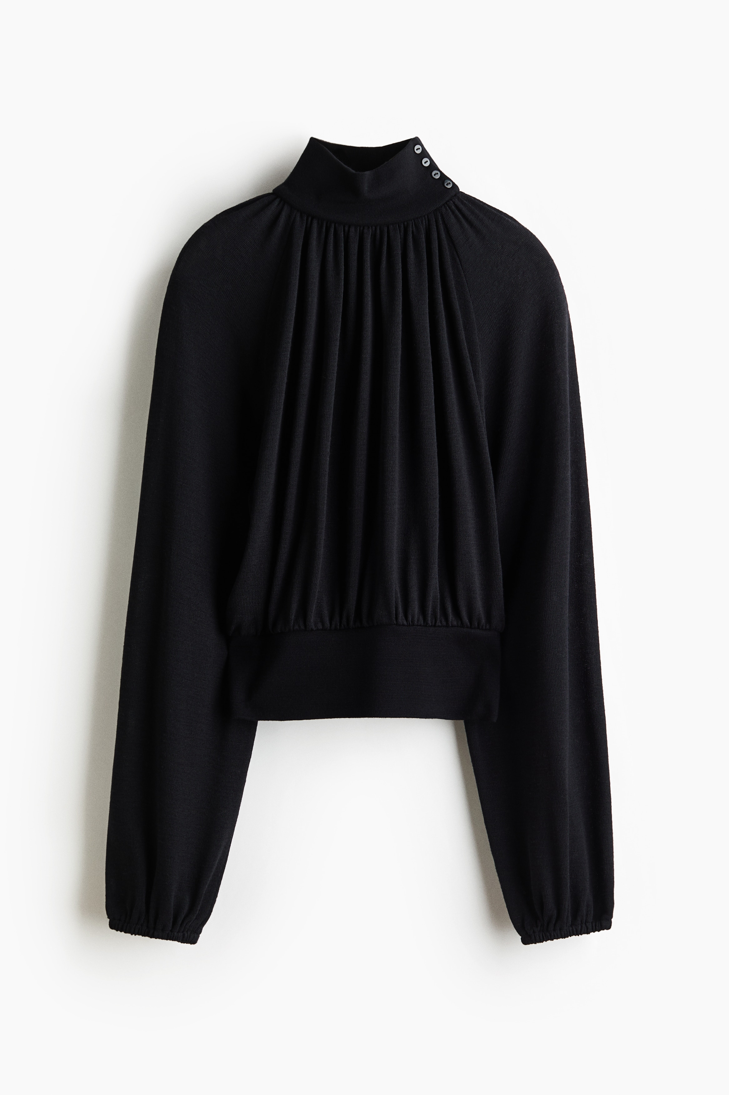 Eine schwarze Bluse mit lockerer Passform, hohem Stehkragen und langen BallonÃ¤rmeln mit gerafften BÃ¼ndchen wird vom Model getragen. Die Bluse wird mit einem breiten weissen GÃ¼rtel, einem kurzen weissen Rock und schwarzen kniehohen Stiefeln kombiniert.