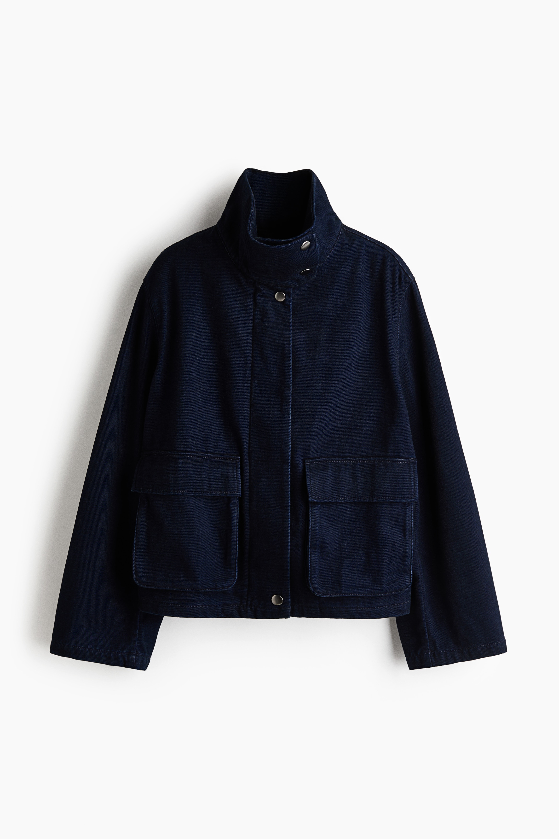 La modelo lleva puesta una chaqueta de sarga azul marino oscuro, con cuello alto tipo chimenea cerrado con broches de presión en tono plateado, hombros caídos y amplios bolsillos de fuelle delanteros con solapa. La modelo aparece de pie con las manos metidas en los bolsillos de unos vaqueros oscuros.
