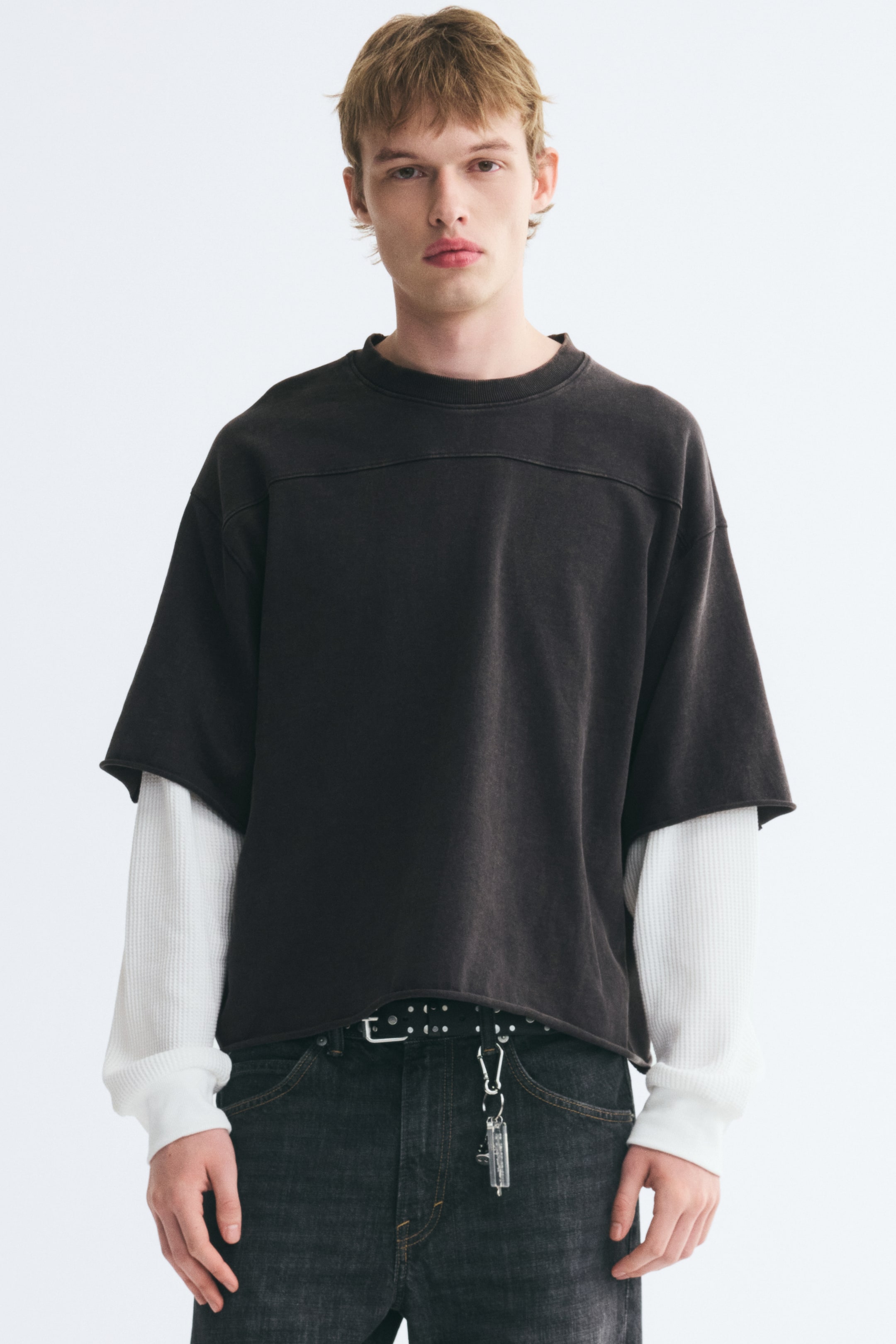 View larger image: A layered, oversized washed-black T-shirt is worn by the model, featuring a front yoke seam, a rib-trimmed crewneck, and raw-edge hems. The design includes integrated long white waffle-knit sleeves extending from the short sleeves and is styled with dark denim jeans.