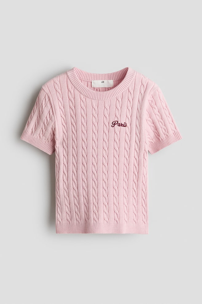 A pink short-sleeved cable-knit top is showcasing its front, featuring a ribbed crew neck and short ribbed sleeves. The word "Paris" is embroidered in a dark script on the front. - 1