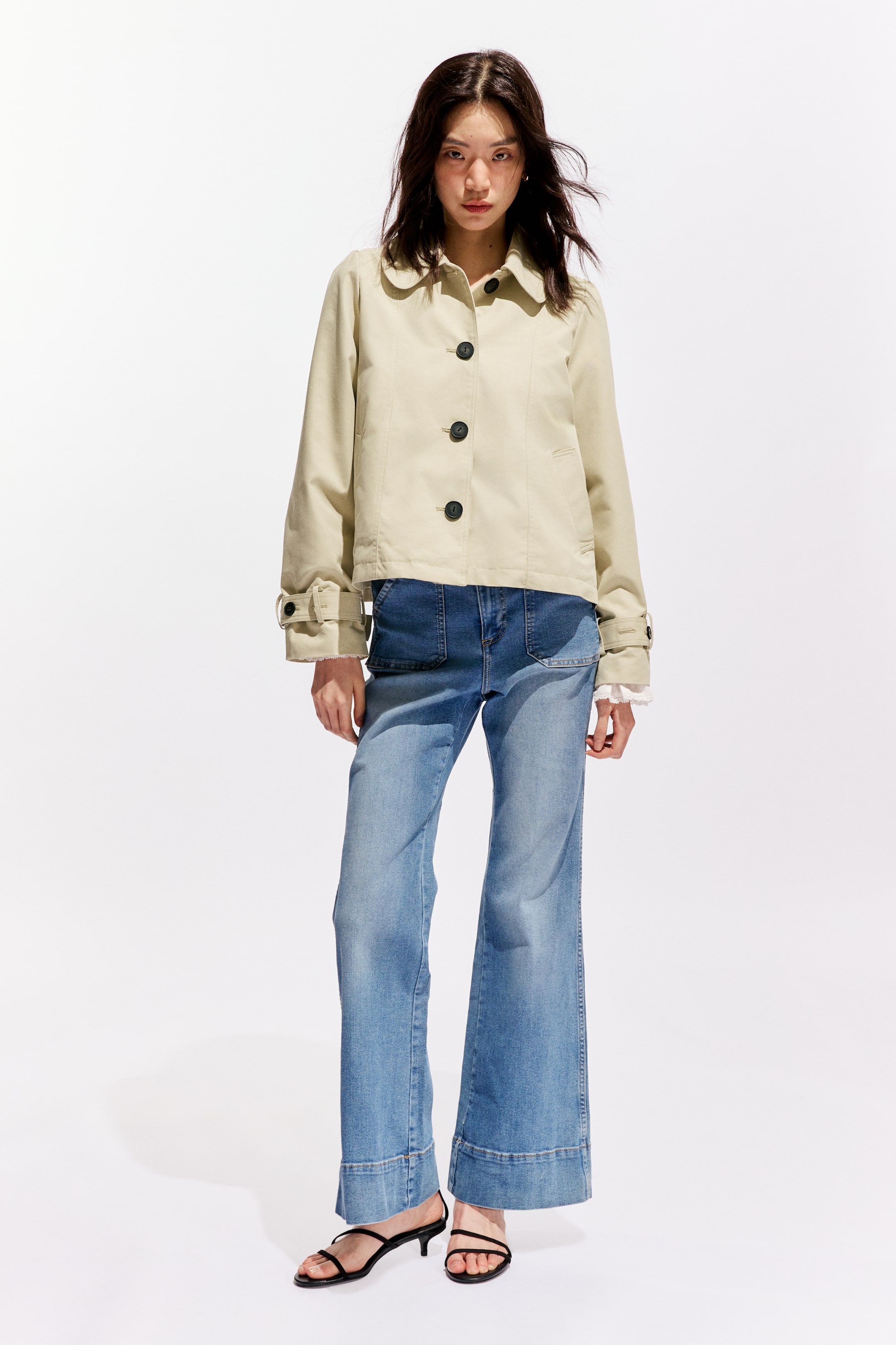 拡大画像を表示: ベージュのクロップジャケットが前を向いて表示されており、尖った襟と前面に並ぶ3つのダークボタンが特徴です。長袖のデザインで、カフスには調整可能なボタンタブが付いており、白いレースのカフスが下から覗いています。