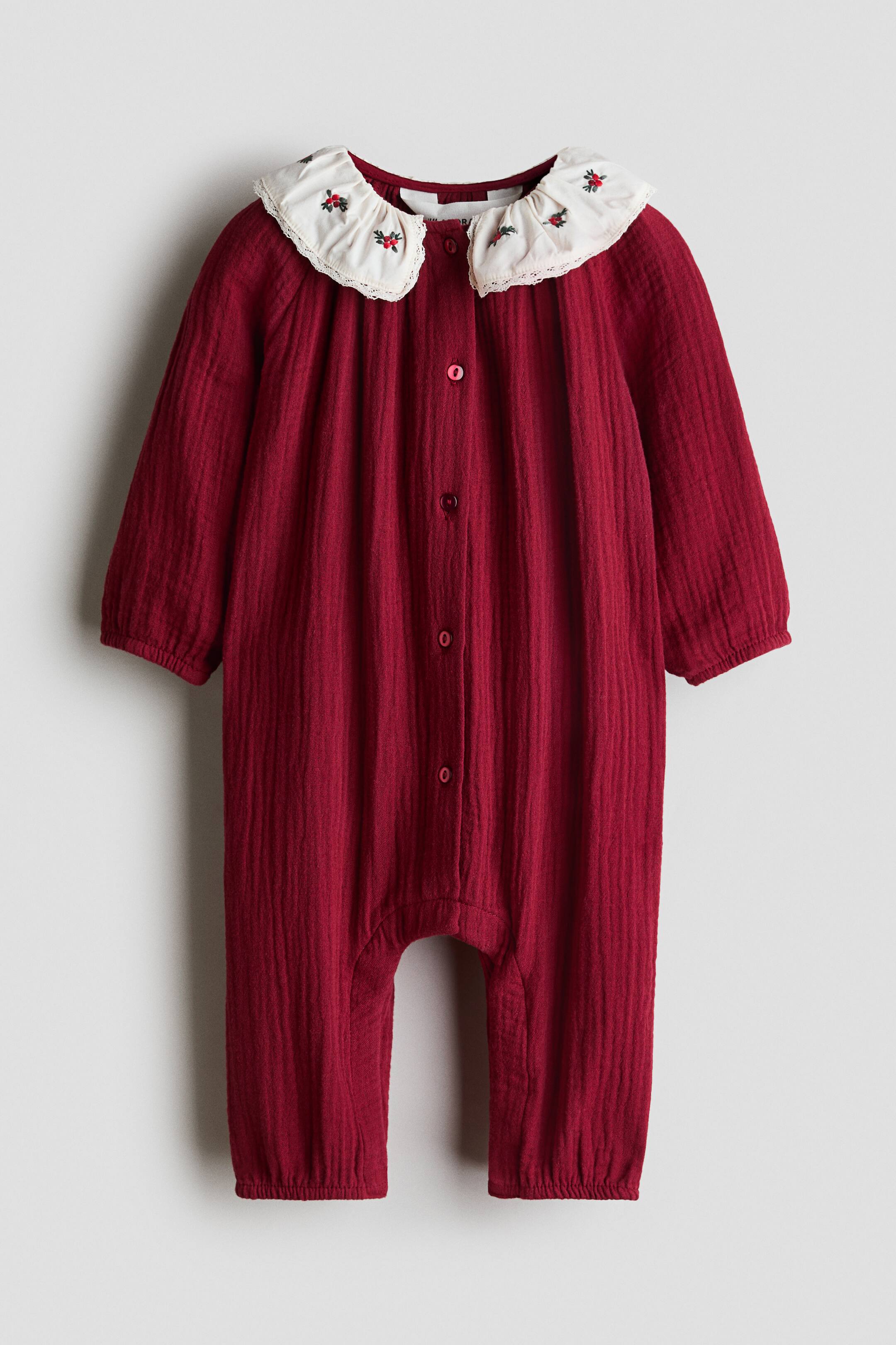Vis større bilde: En dyp rød babyheldress i musselin vises forfra, med lange ermer, knappelukking foran og elastiske mansjetter og avslutninger. Den har en kontrasterende off-white krage med bølgekant, dekorert med små broderte røde blomsterdetaljer og delikat blondekant.
