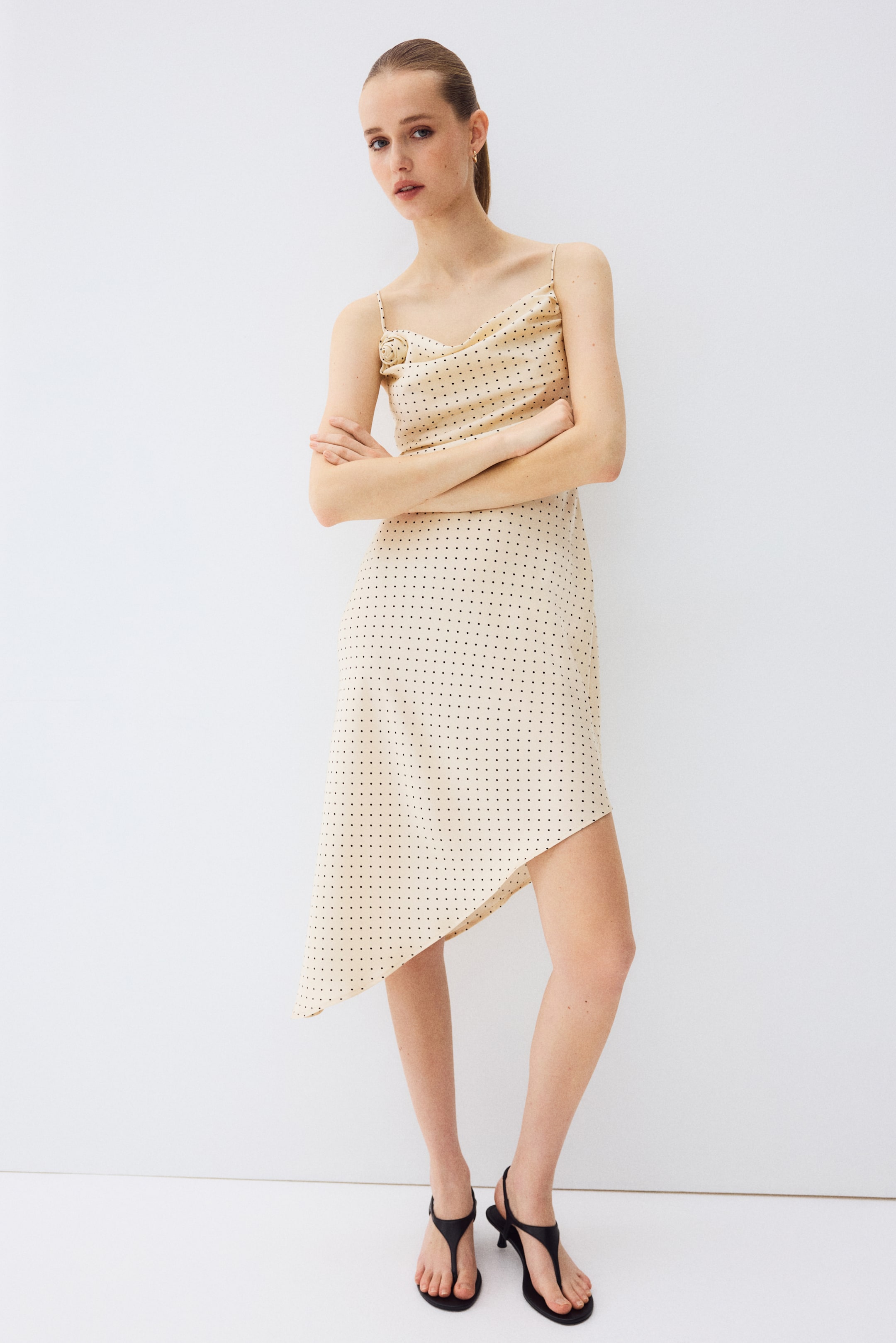 Ampliar la imagen: Un vestido de tirantes de color beige claro con lunares negros presenta finos tirantes de espagueti y un escote drapeado en forma de cowl, con un adorno de rosetón en el busto izquierdo. El vestido luce un dobladillo asimétrico, visto desde el frente.