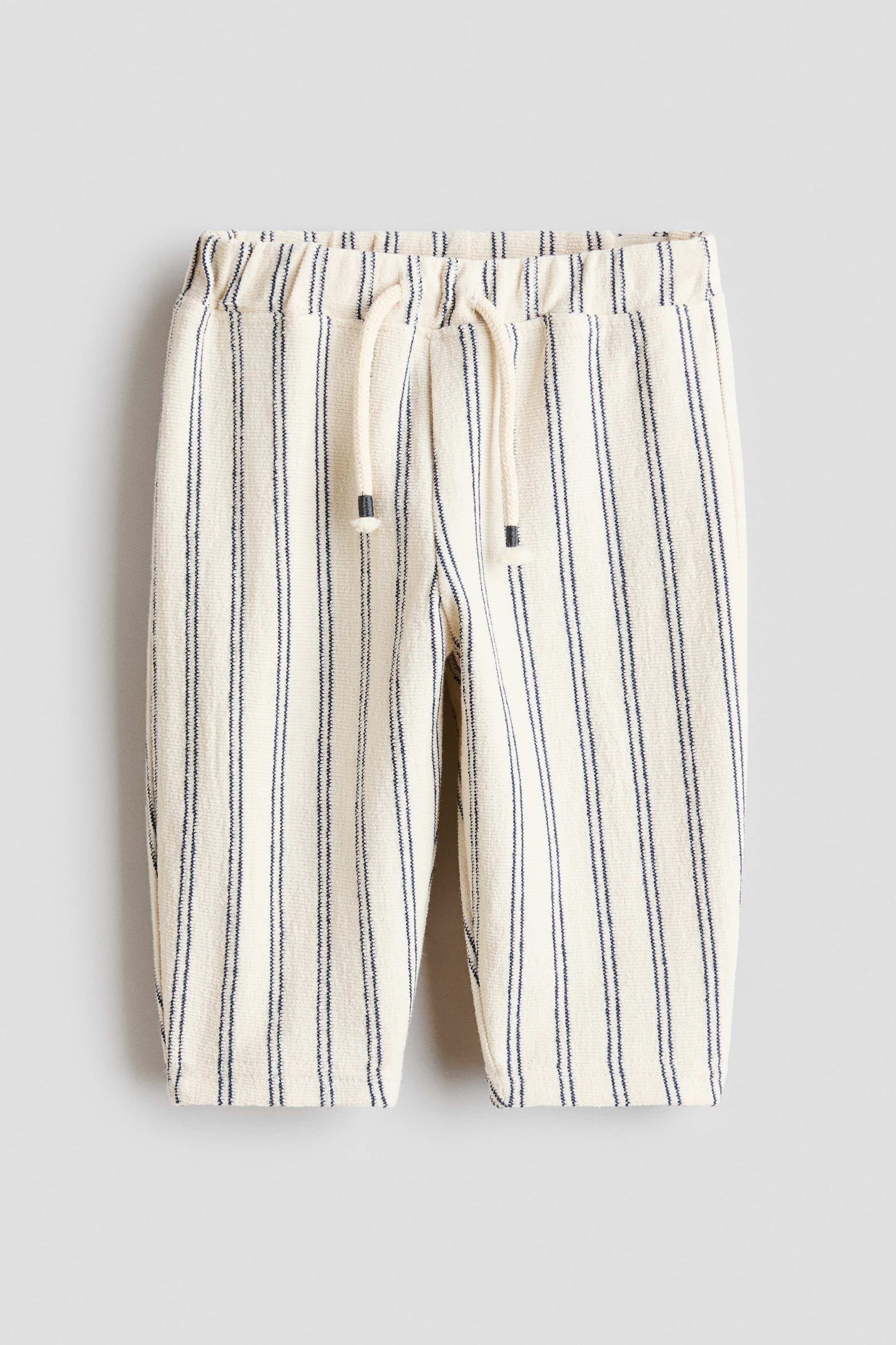 הצגת תמונה גדולה יותר: המכנסיים הללו מוצגים מהחזית, עם בד ג'רזי קרם רך ומרקם עם פסים אנכיים כחולים כהים. הם מציגים מותן אלסטית מתכווננת עם חוט קשירה דקורטיבי בצבע קרם המסתיים בקצוות כהים.