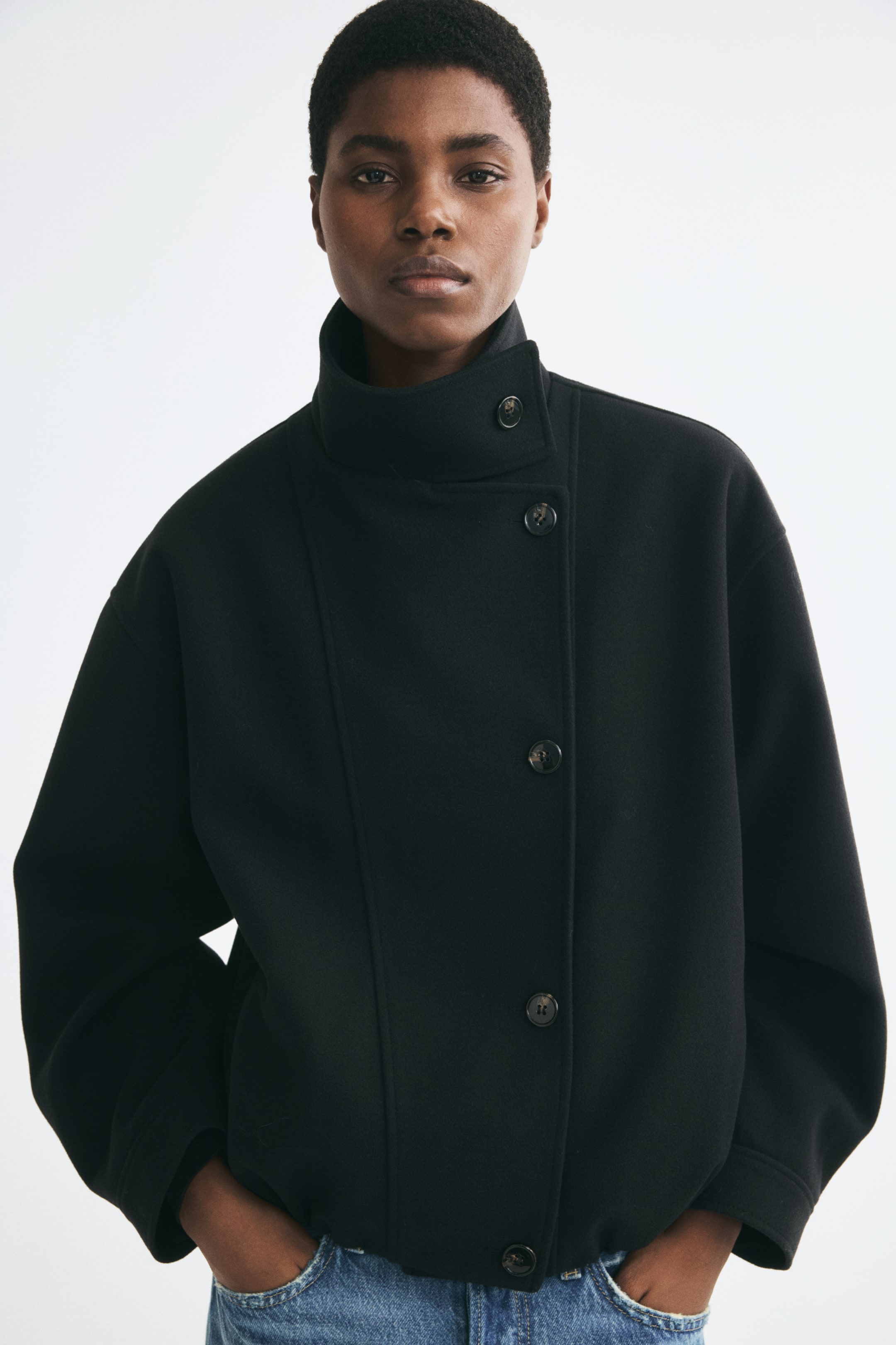 Agrandir l'image: Une veste noire oversize en tissu feutré doux est portée par le mannequin, avec un col cheminée boutonné et une fermeture à boutons décalée sur le devant. Le mannequin se tient debout, les mains glissées dans les poches d’un jean en denim bleu visible sous l’ourlet de la veste.