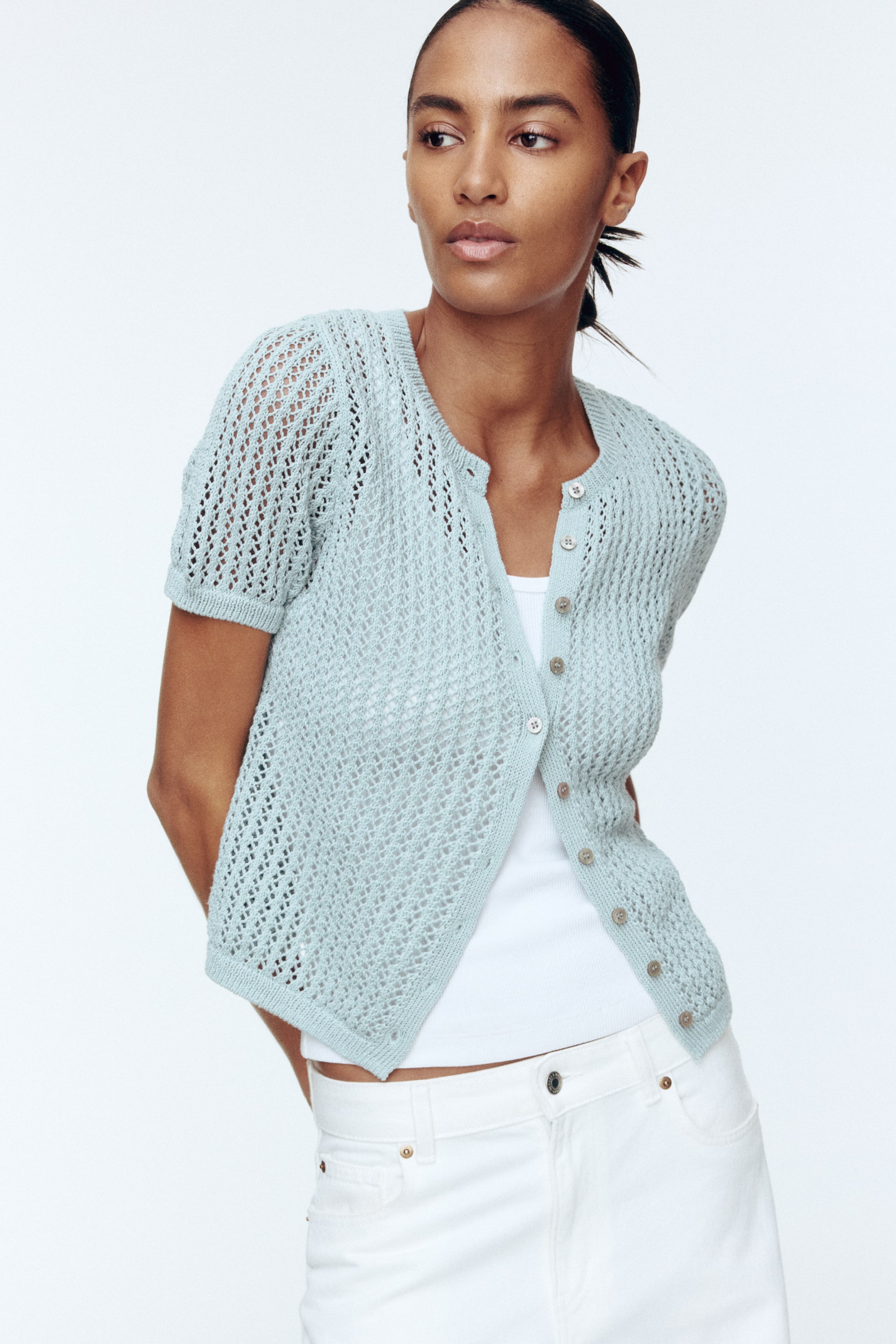 View larger image: A light blue, short-sleeved, open-knit cardigan is being displayed from the front, angled slightly right, featuring a round neckline and a button-front closure with small light-colored buttons. It is showing ribbed cuffs and a ribbed hem, and is being worn unbuttoned over a white tank top.