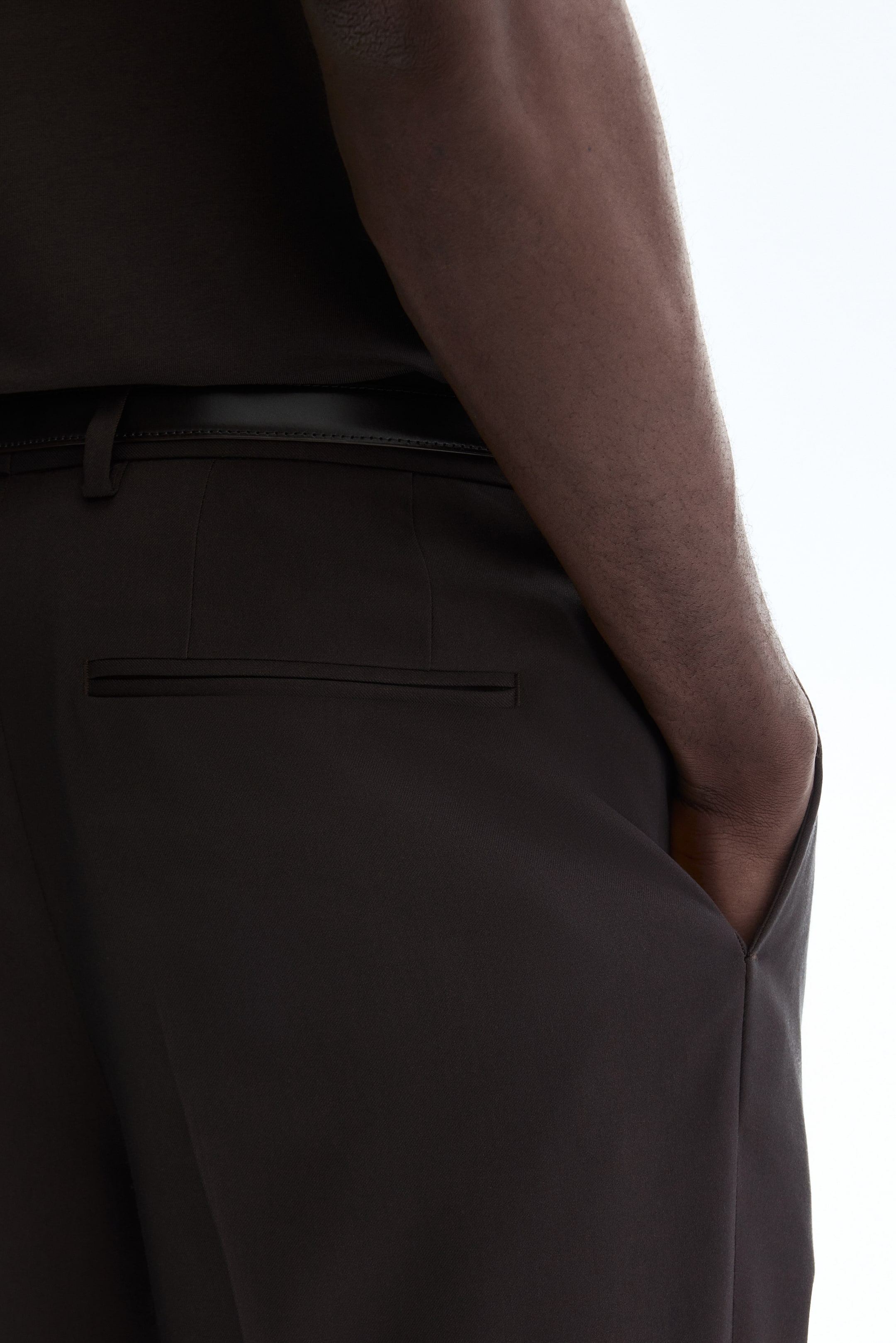 Ampliar la imagen: El modelo lleva puestos pantalones de traje en tela tejida. Los pantalones cuentan con un bolsillo trasero de ojal y trabillas para cinturón, y el modelo coloca su mano en un bolsillo lateral mientras usa un cinturón oscuro.