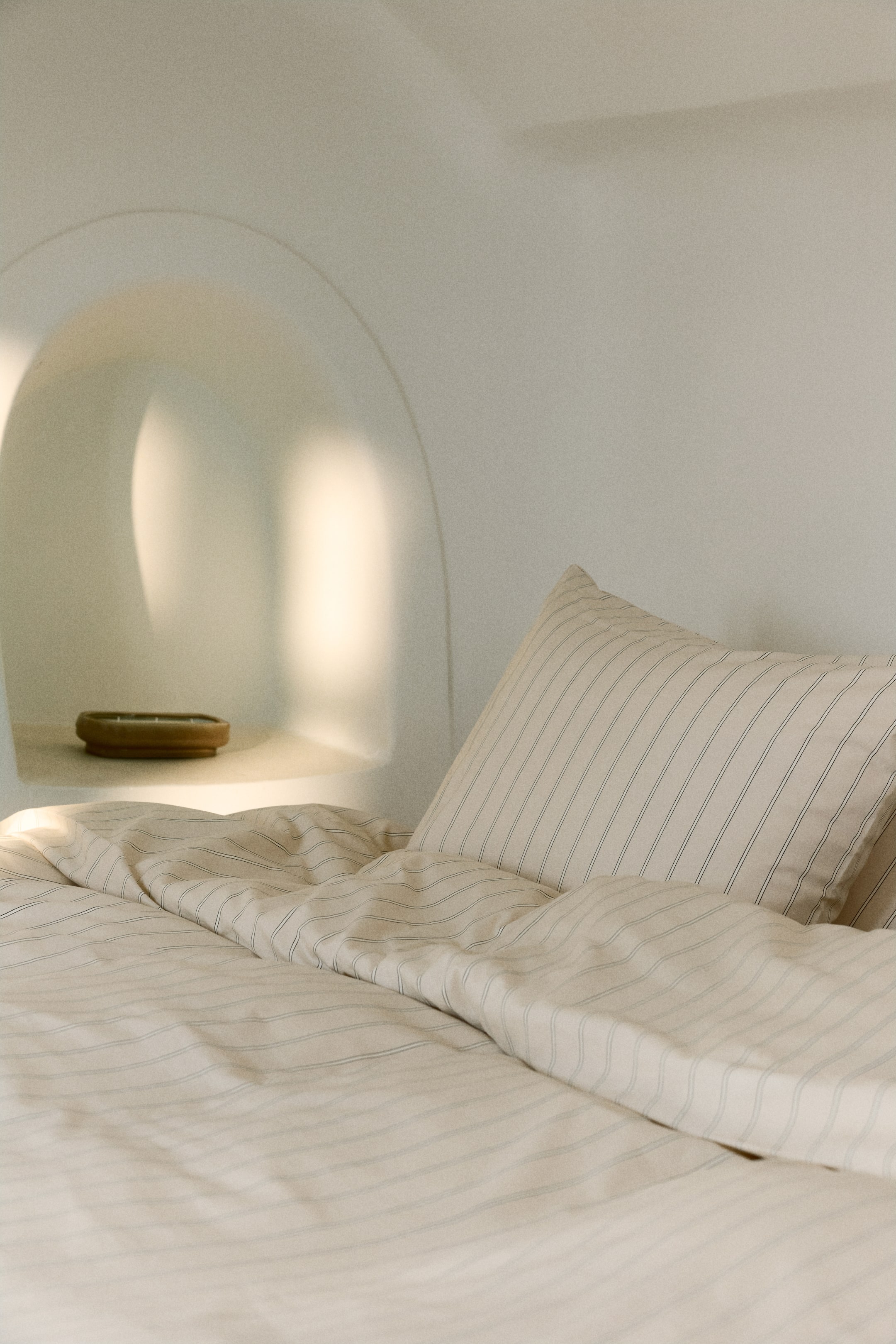 View larger image: A light cream bed linen set, featuring thin dark pinstripes, is depicting a pillowcase standing upright and a duvet cover draped over a bed. The pillowcase is angled slightly to the right, and the duvet cover is exhibiting soft folds and creases.