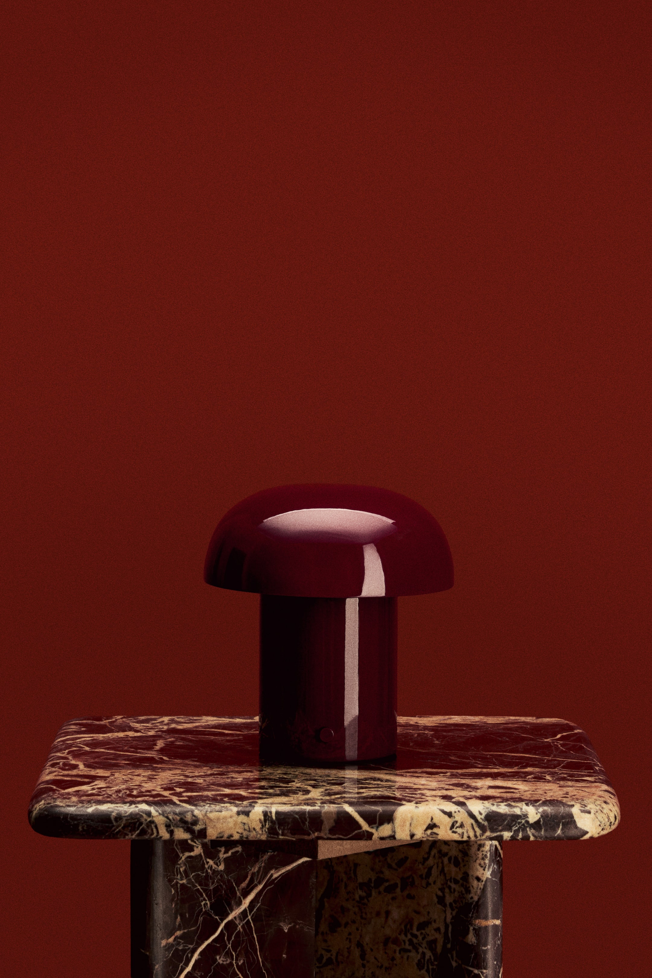 View larger image: The rechargeable table lamp showcases a glossy, dark red metal construction. It features a rounded, mushroom-inspired design with a domed cap and a cylindrical base, positioned front-facing.
