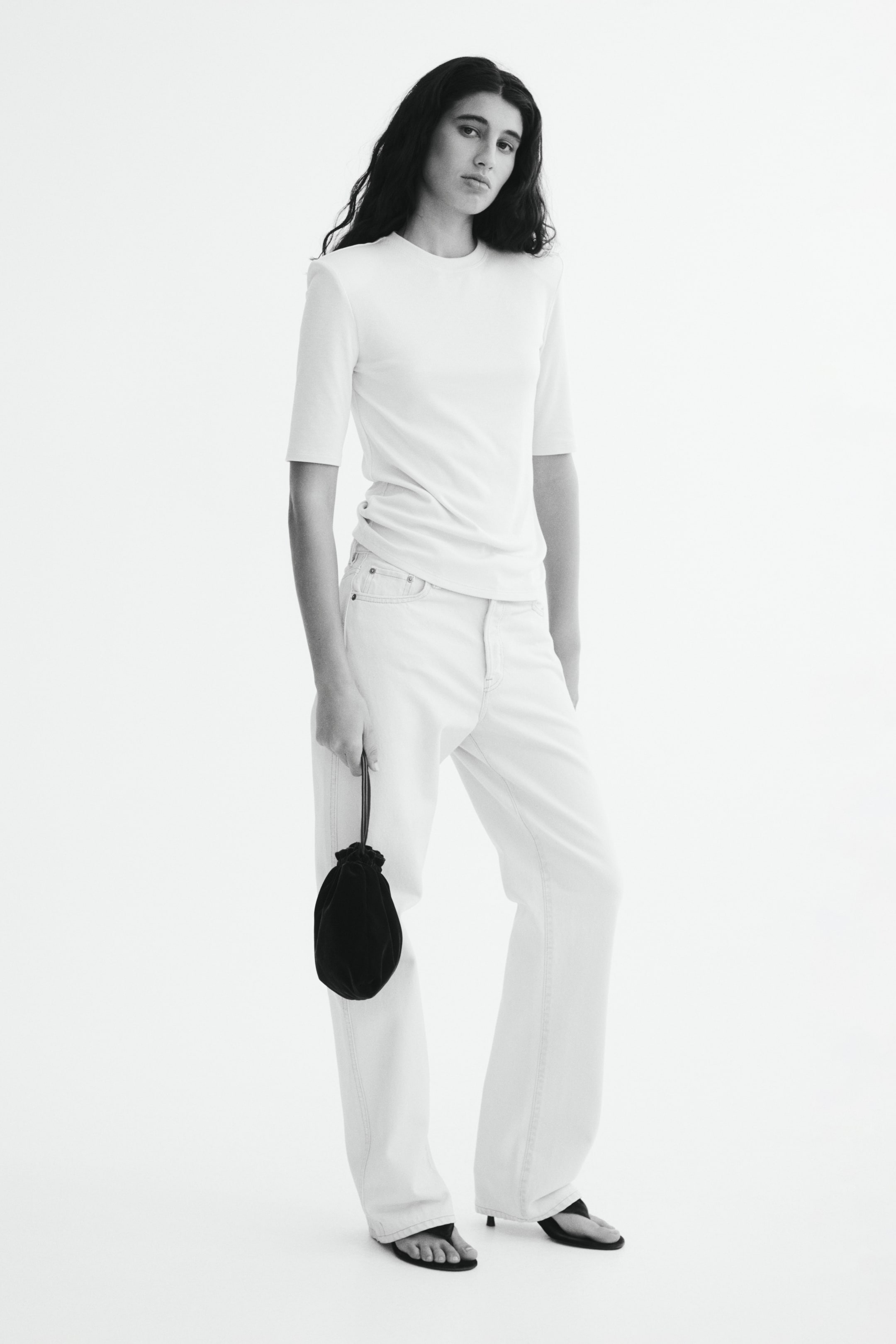 View larger image: The model is wearing a fitted ribbed jersey tee with a crew neckline, structured shoulder pads, and a straight hem. It is styled with light-wash jeans, and the model is holding a small black drawstring bag.
