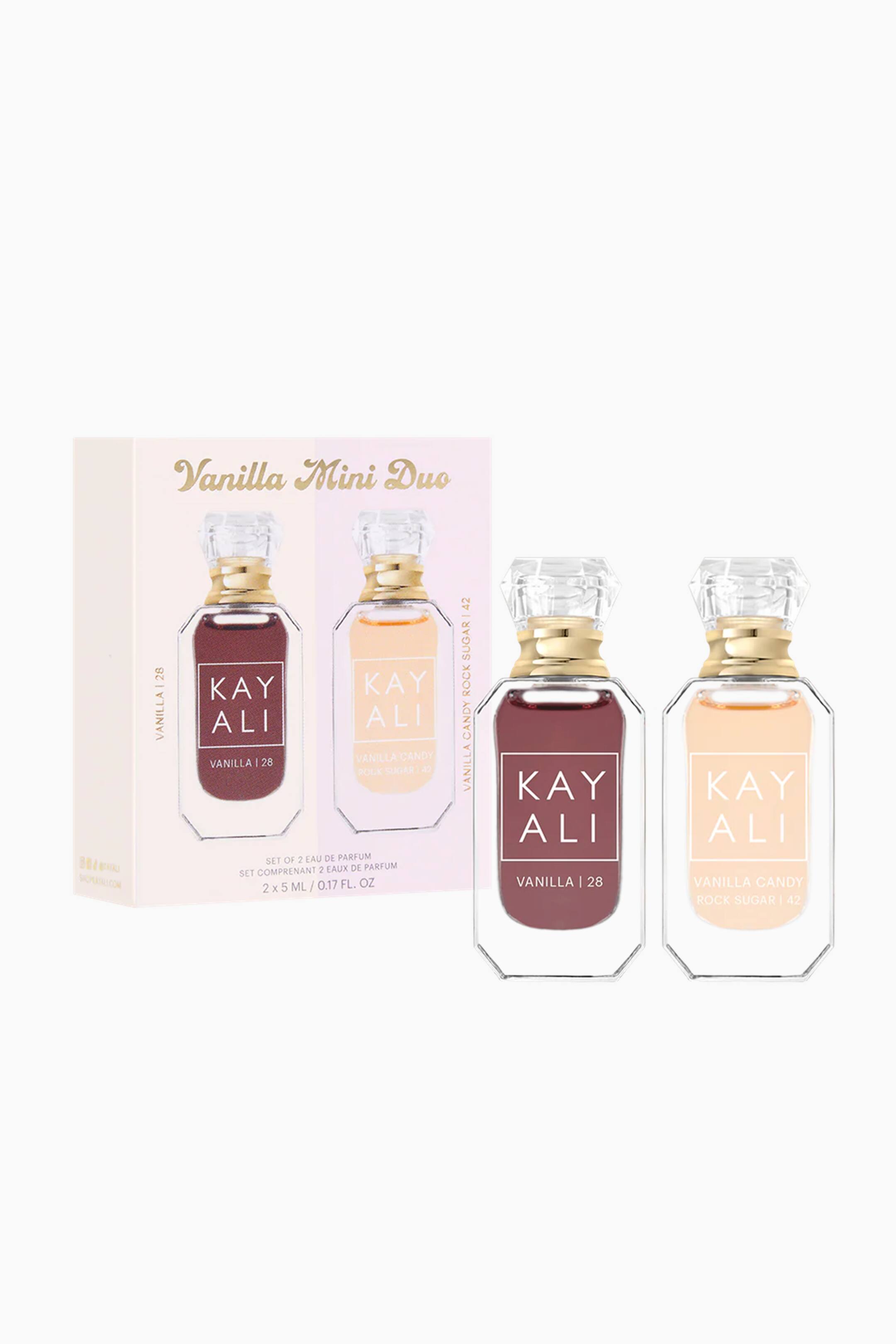 Vis større bilde: Et 'Vanilla Mini Duo' parfyme sett vises, med en lys rosa og beige boks og to mini parfymeflasker, alle vendt litt fremover og til høyre. Boksen viser utskjæringer for de to flaskene, som indikerer 'SET OF 2 EAU DE PARFUM 2 x 5 ML / 0.17 FL. OZ.' De klare glassflaskene, sekskantede ved basen, har gullfargede halser og klare fasetterte lokk; den ene inneholder mørk rødbrun væske merket 'KAY ALI VANILLA | 28,' og den andre inneholder lys ferskenfarget væske merket 'KAY ALI VANILLA CANDY ROCK SUGAR | 42.'