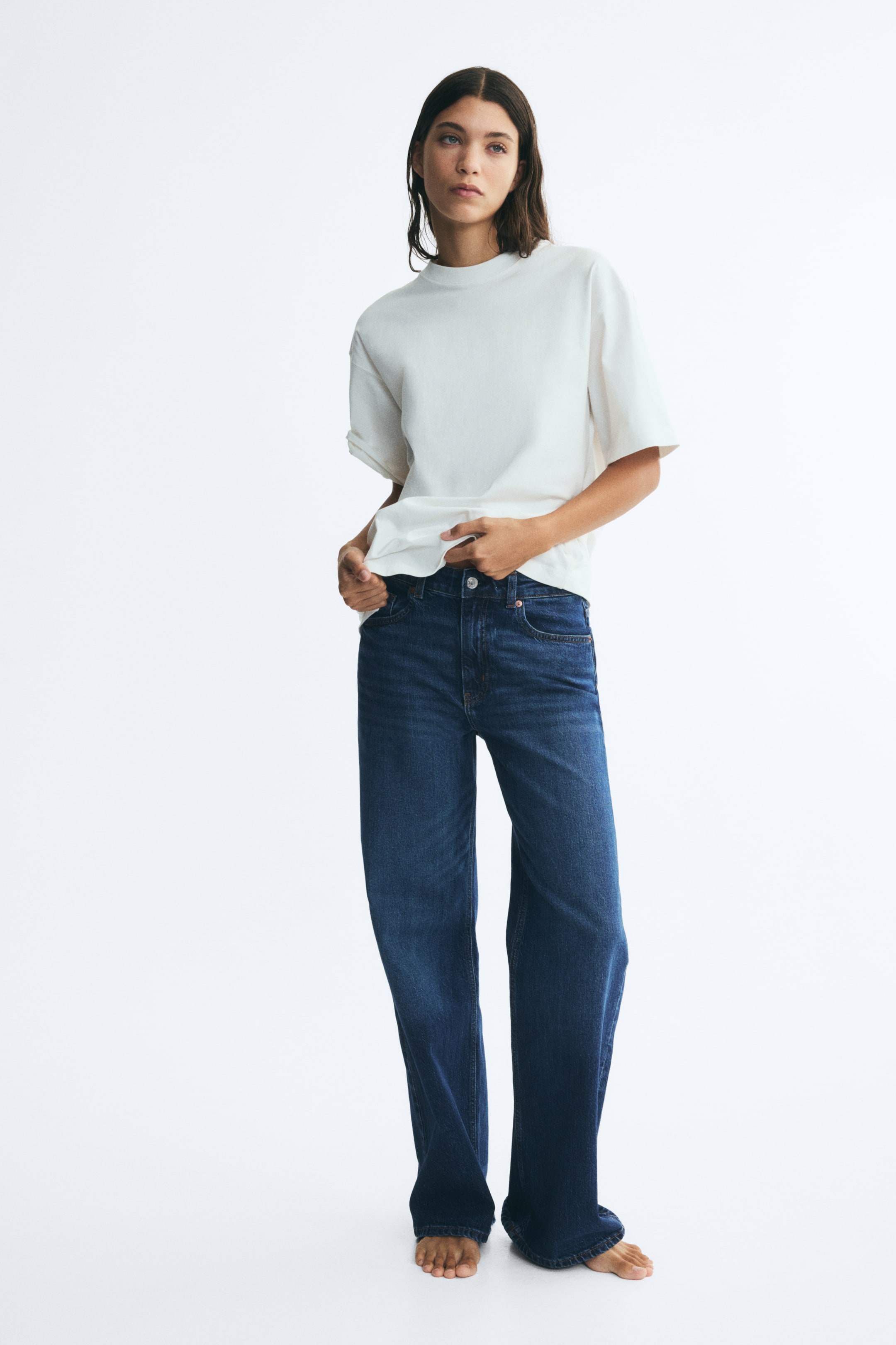 Visualizza immagine più grande: La modella indossa jeans blu scuro a vita alta e gamba larga, sollevando l’orlo di una t-shirt bianca per mettere in evidenza la vita e la chiusura con bottone. I jeans presentano un classico design a cinque tasche e una vestibilità morbida che arriva fino ai piedi.