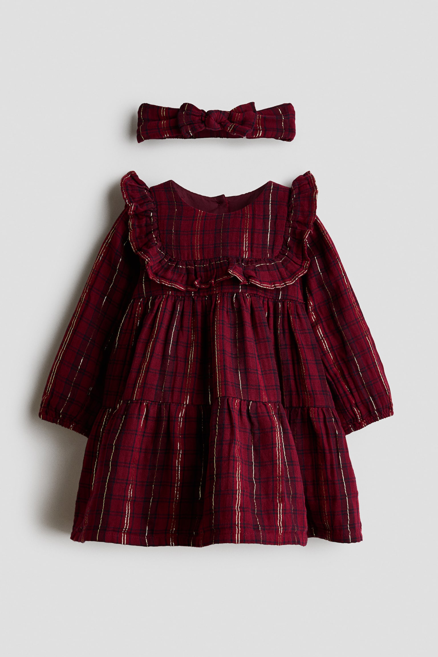 A baby dress and matching headband set is shown from the front, featuring a dark red plaid pattern with metallic gold vertical stripes. The long-sleeved dress features a ruffled bib collar, a tiered gathered skirt, and elasticated cuffs, while the matching fabric headband showcases a knotted bow.