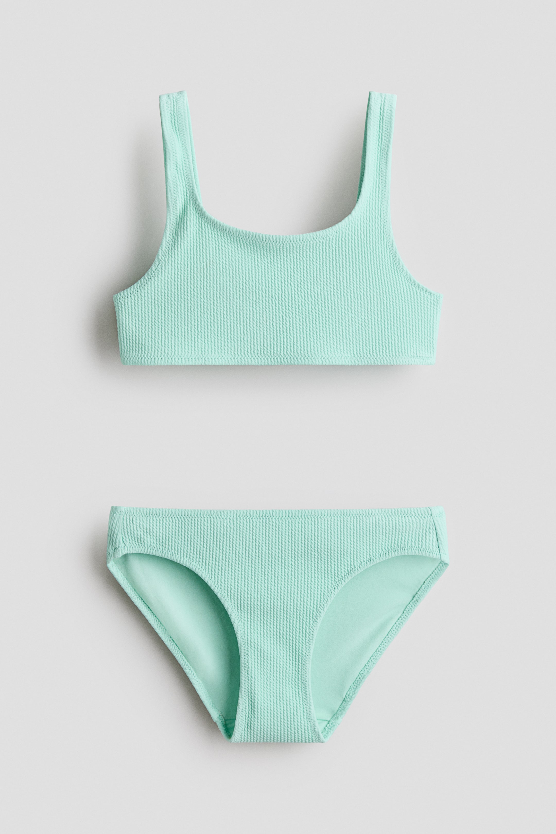 Größeres Bild anzeigen: Ein vollständig gefüttertes Bikini-Set präsentiert ein Oberteil und passende Unterteile, beide in einem hellgrünen strukturierten Stoff. Das Oberteil verfügt über breite Schulterträger und einen quadratischen Ausschnitt, während die Unterteile im klassischen Slip-Stil gehalten sind.