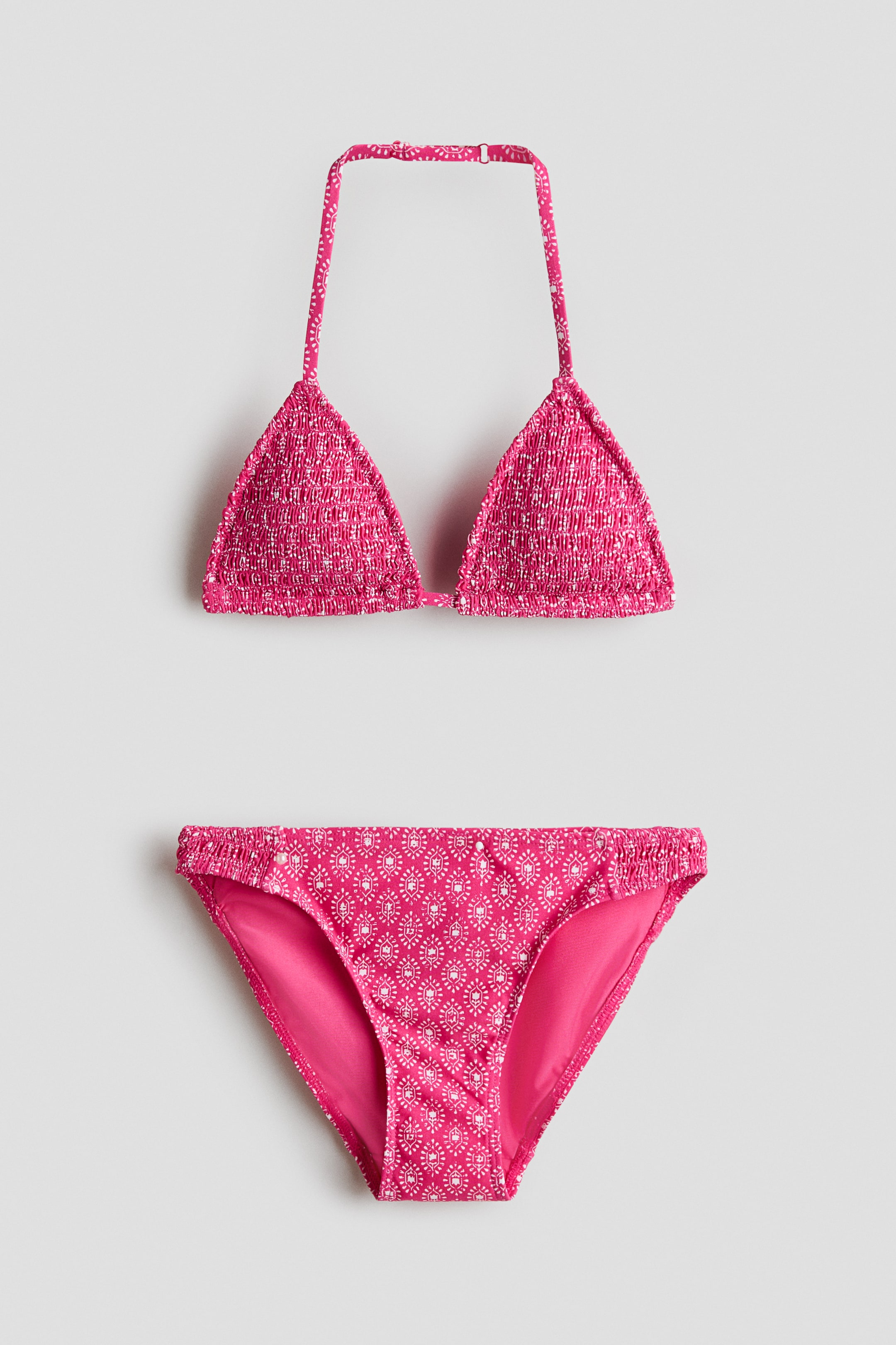 View larger image: A pink bikini set with a white geometric pattern is shown from the front. The smocked triangle bikini top features a halterneck and a plastic fastener at the back, while the matching bottoms display smocked sections at the sides.