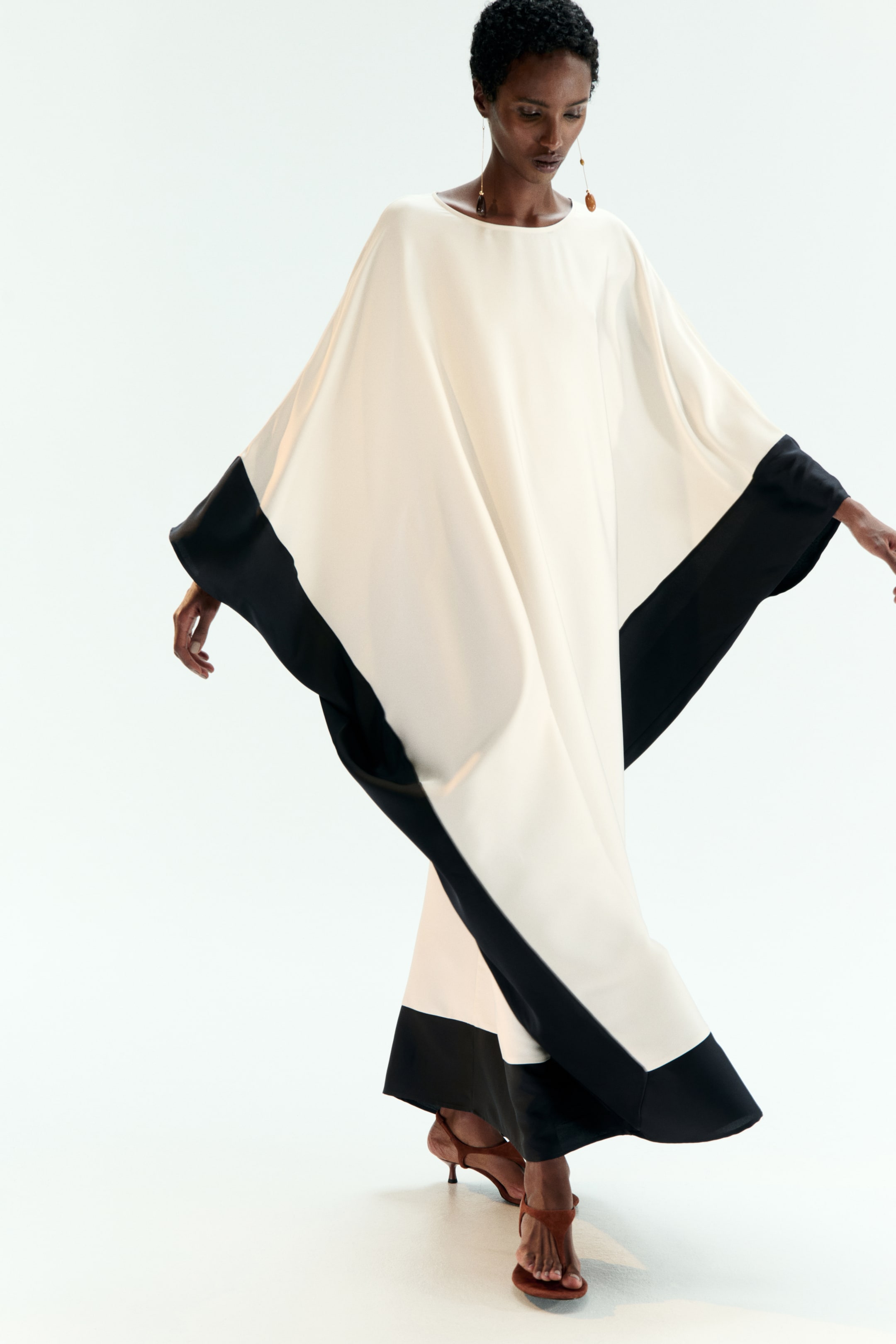 View larger image: A cream kaftan dress is featuring a wide, flowing silhouette with a round neckline and broad black panels accenting the lower sleeves and hem.