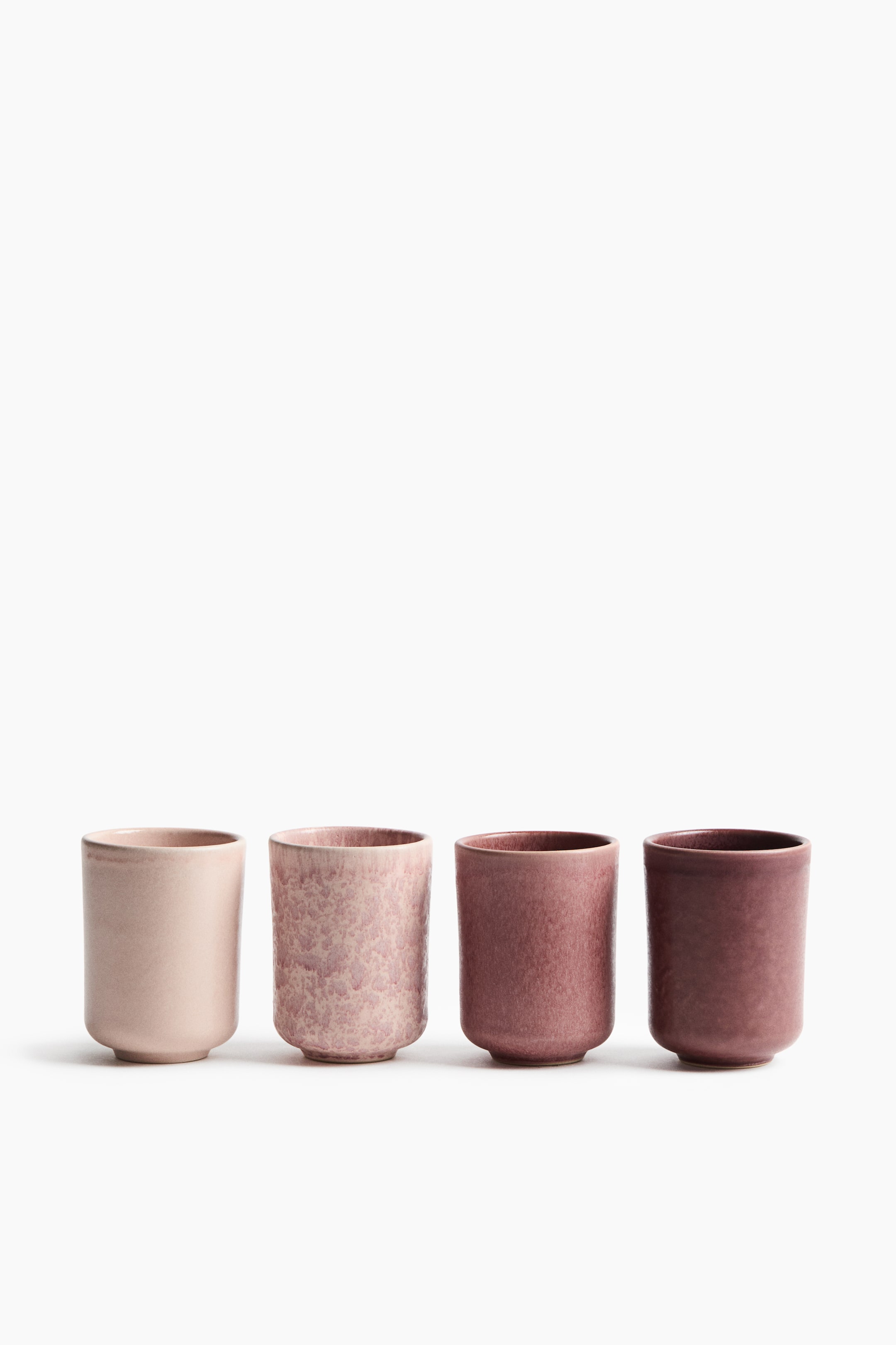 Agrandir l'image: Quatre tasses en grès sans anse sont alignées face à l’avant, affichant un dégradé d’émaux rose et mauve aux finitions texturées uniques. Les tasses passent d’une teinte rose pâle lisse à gauche, à un rose moucheté parsemé de taches, puis à un rose poudré texturé, pour finir par un émail prune profond à l’extrême droite.