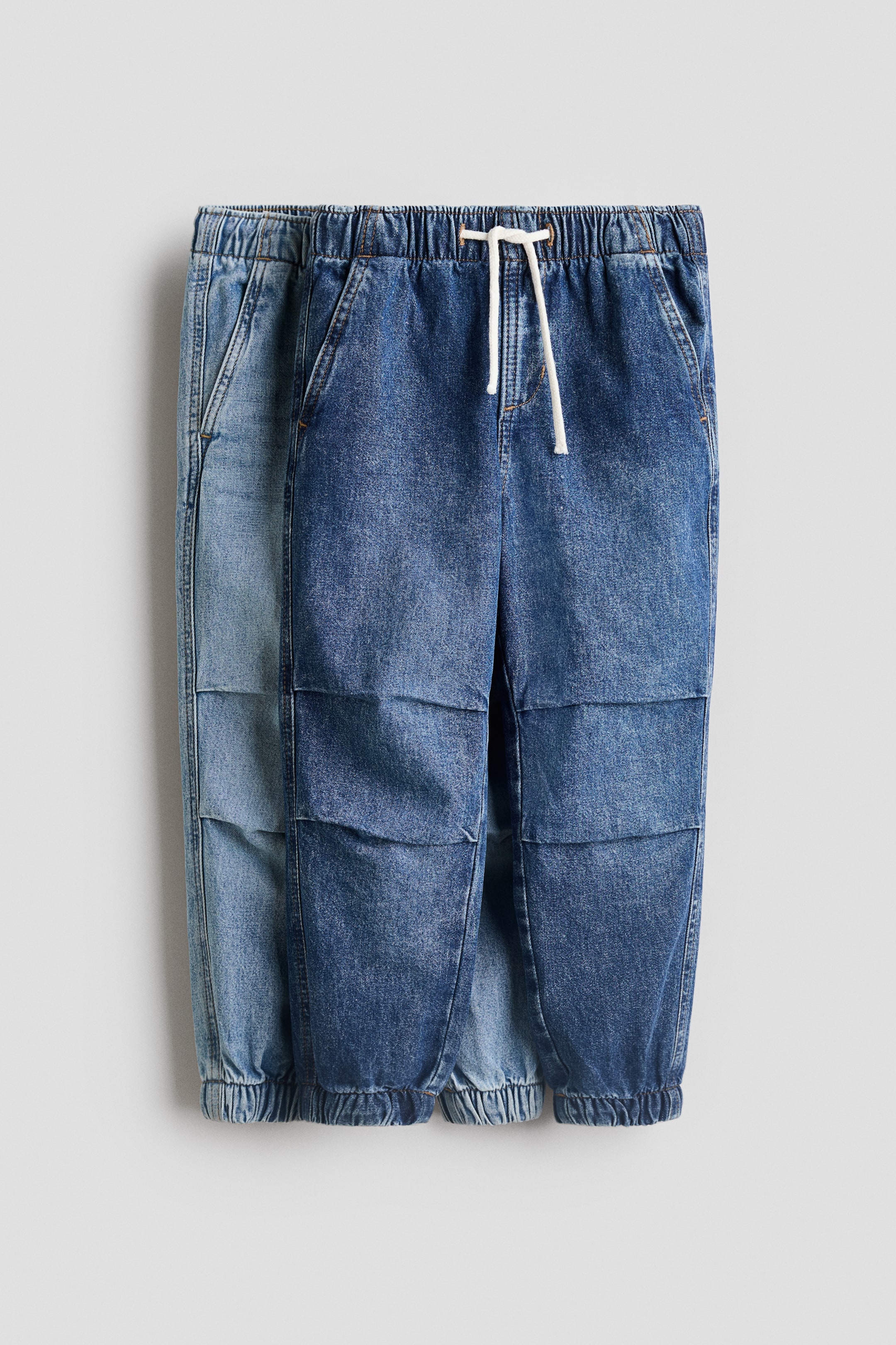 View larger image: Denim jogger pants are presented FRONT, featuring an elasticated waistband with a white drawstring and elasticated cuffs. The medium blue pair is displaying a paneled design on the legs, while a lighter blue pair is partially visible behind it.