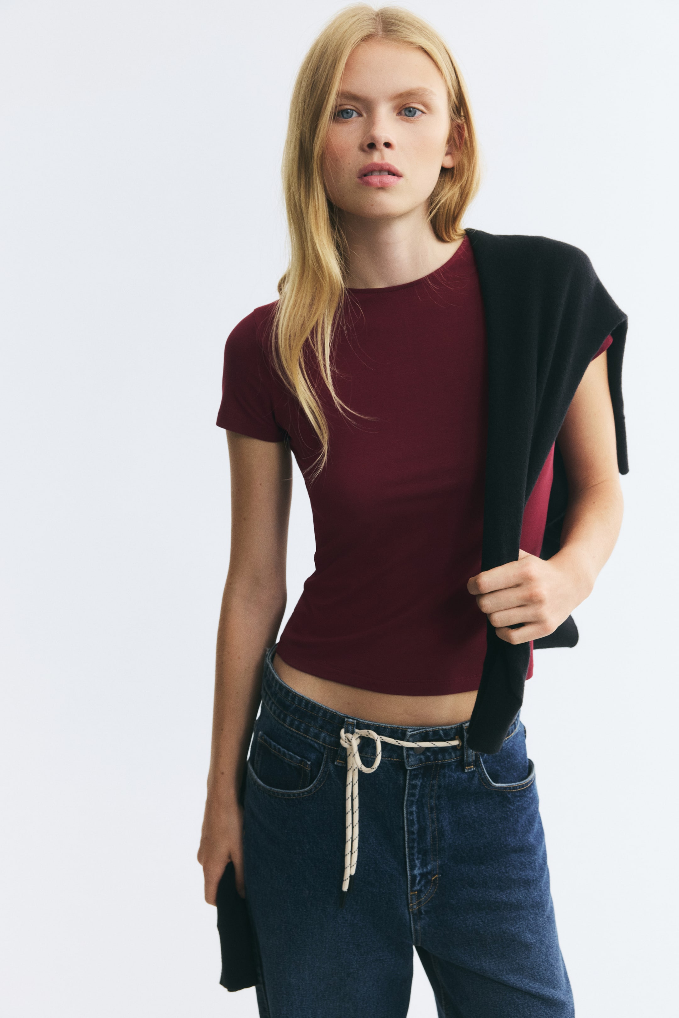View larger image: A burgundy fitted tee with a crew neck and short sleeves is being worn by the model. The model is holding a black sweater draped over one shoulder, styled with blue jeans.