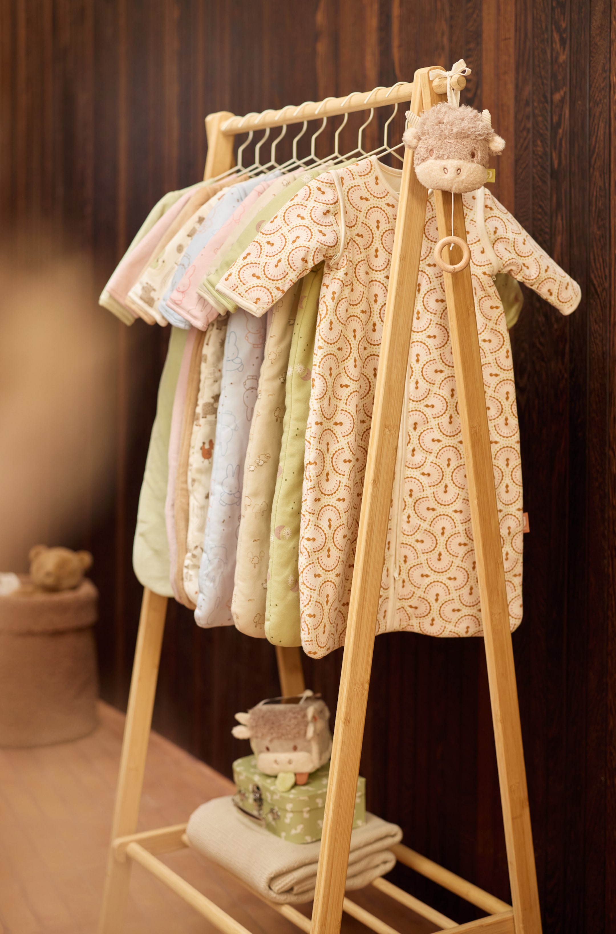 Grotere afbeelding bekijken: Verschillende baby slaapzakken in diverse pastelkleurige tinten en patronen hangen aan draadkleerhangers aan een houten kledingrek. De voorste slaapzak toont een herhalend boogpatroon in beige en bruin, met lange mouwen en een doorlopende ritssluiting aan de voorkant.