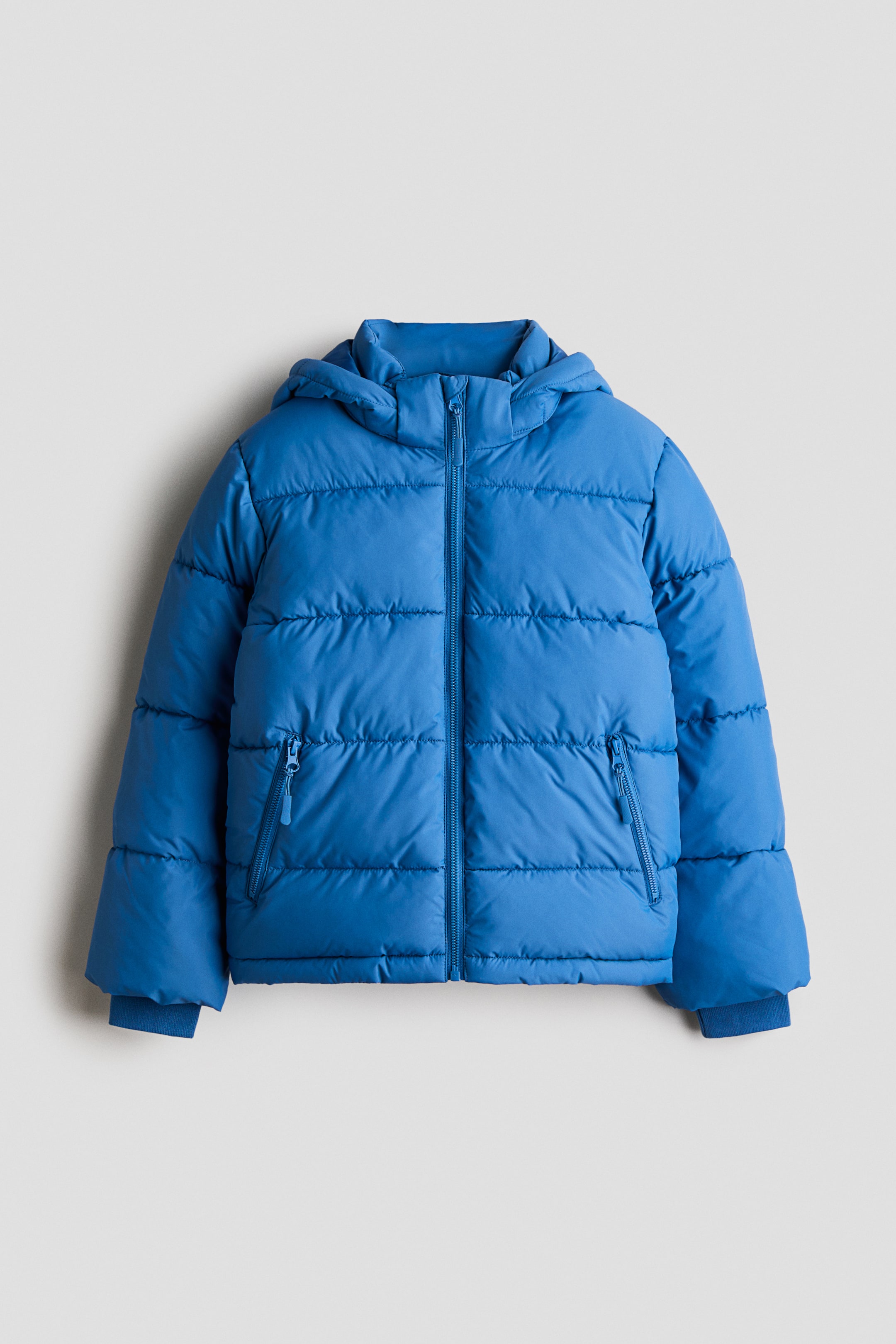 Ver imagen más grande: Una chaqueta acolchada azul se presenta de frente, con capucha incorporada y cierre de cremallera completo. Muestra un acolchado horizontal, dos bolsillos laterales con cremallera y puños acanalados en las mangas.