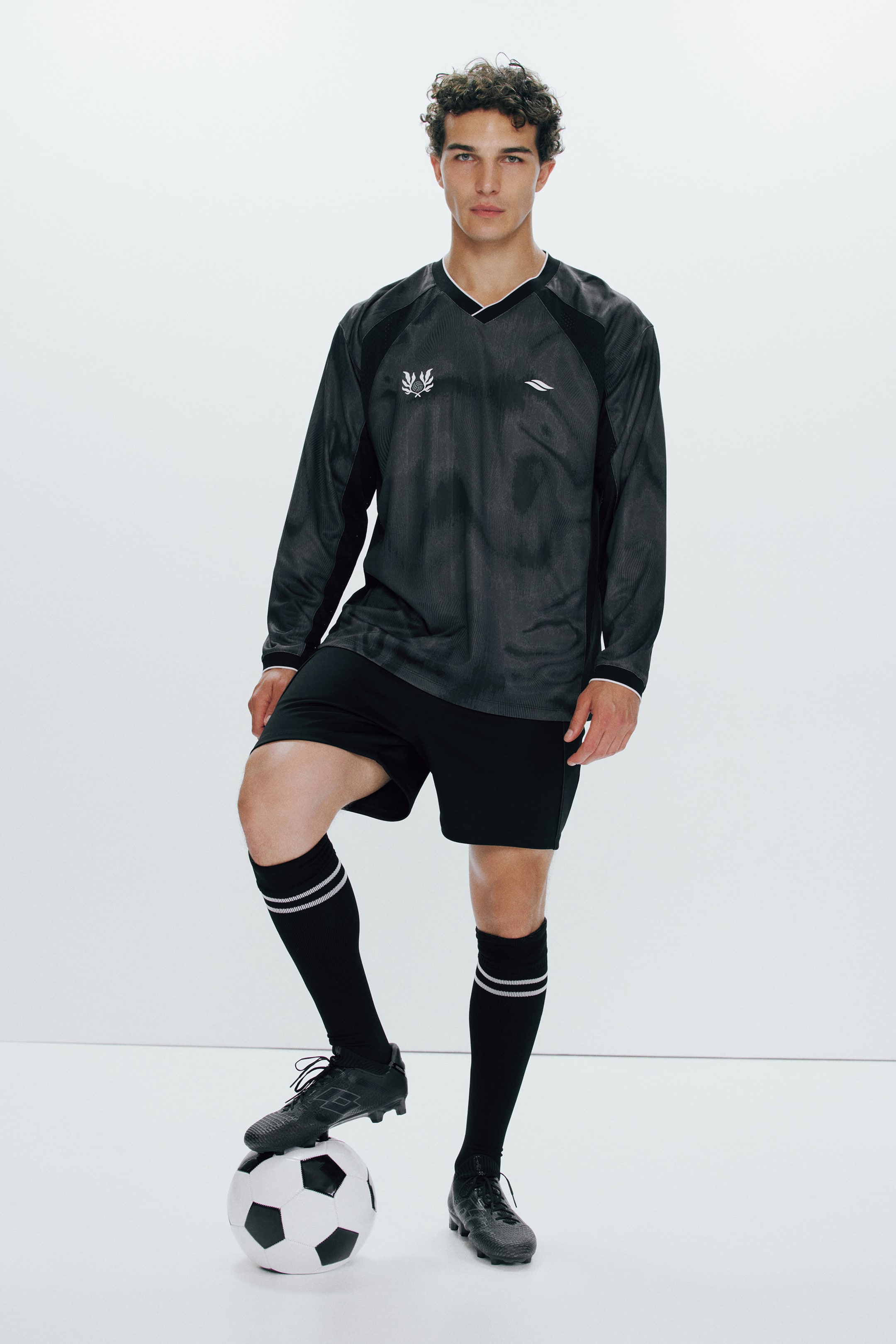 Visualizza l'immagine ingrandita: La modella indossa una maglia da calcio a vestibilità ampia con DryMove?, caratterizzata da un tessuto grigio scuro con motivo sottile, pannelli laterali in rete nera, scollo a V con bordi bianchi e loghi bianchi sul petto. La modella indossa anche pantaloncini neri e calzettoni da calcio, e appoggia un piede su un pallone.