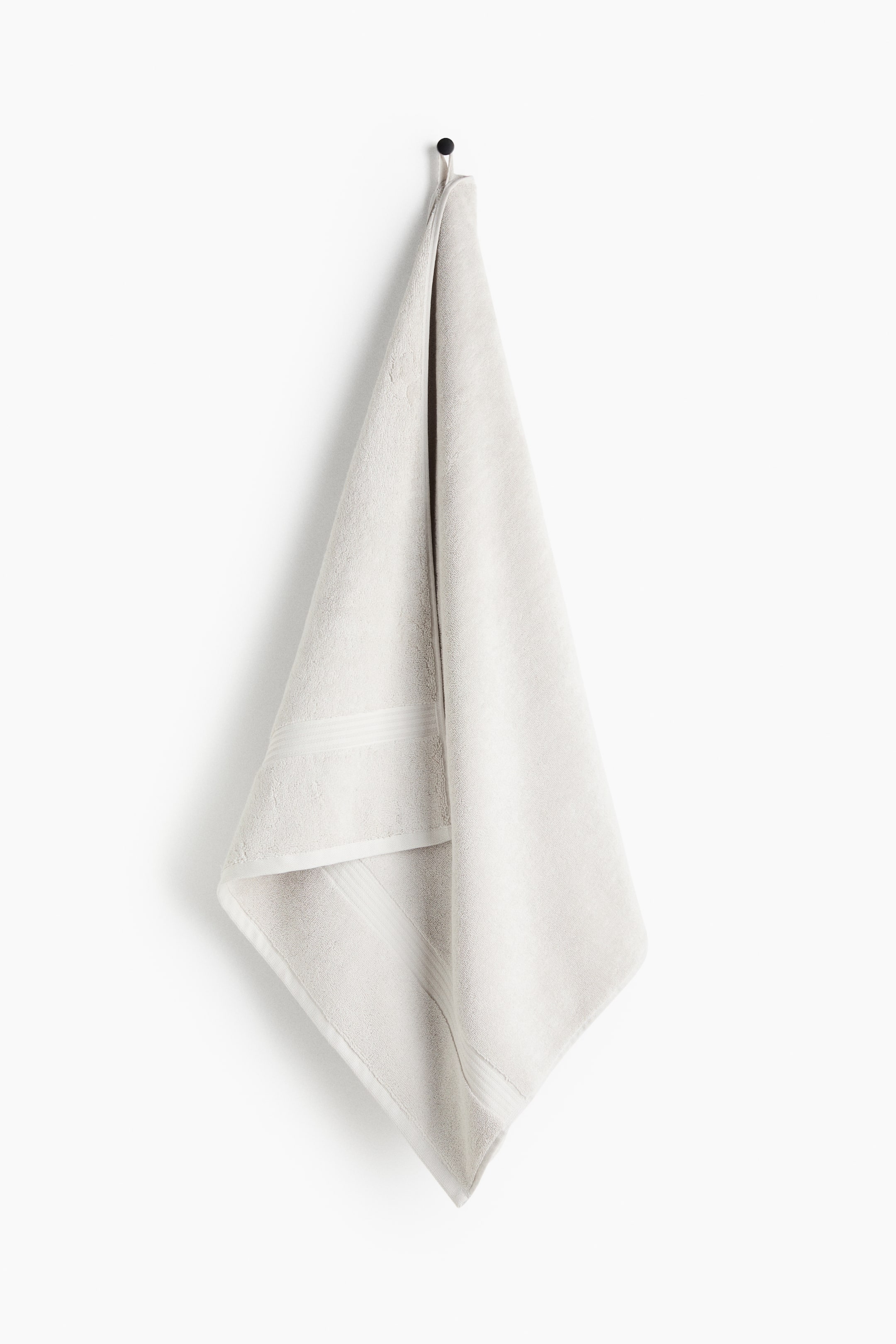 View larger image: A light beige bath towel is hanging, showcasing its cotton terry texture with a subtle sheen and a woven trim along its short sides. A visible loop is holding it from a hook at one long side.