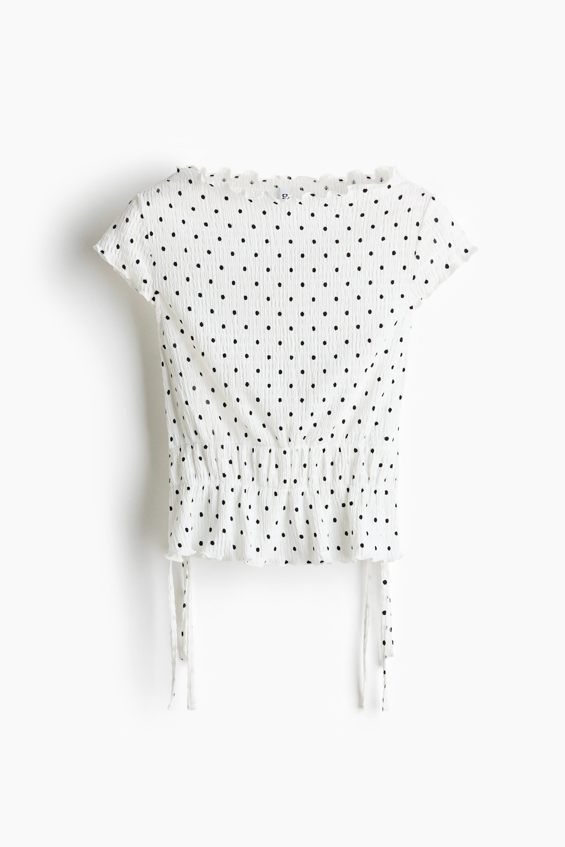 View larger image: A white top with small black polka dots shown from the back, featuring textured fabric all over, a ruffled neckline, and ruffled cap sleeves. It cinches at the waist with a smocked band, from which two white tie strings hang.