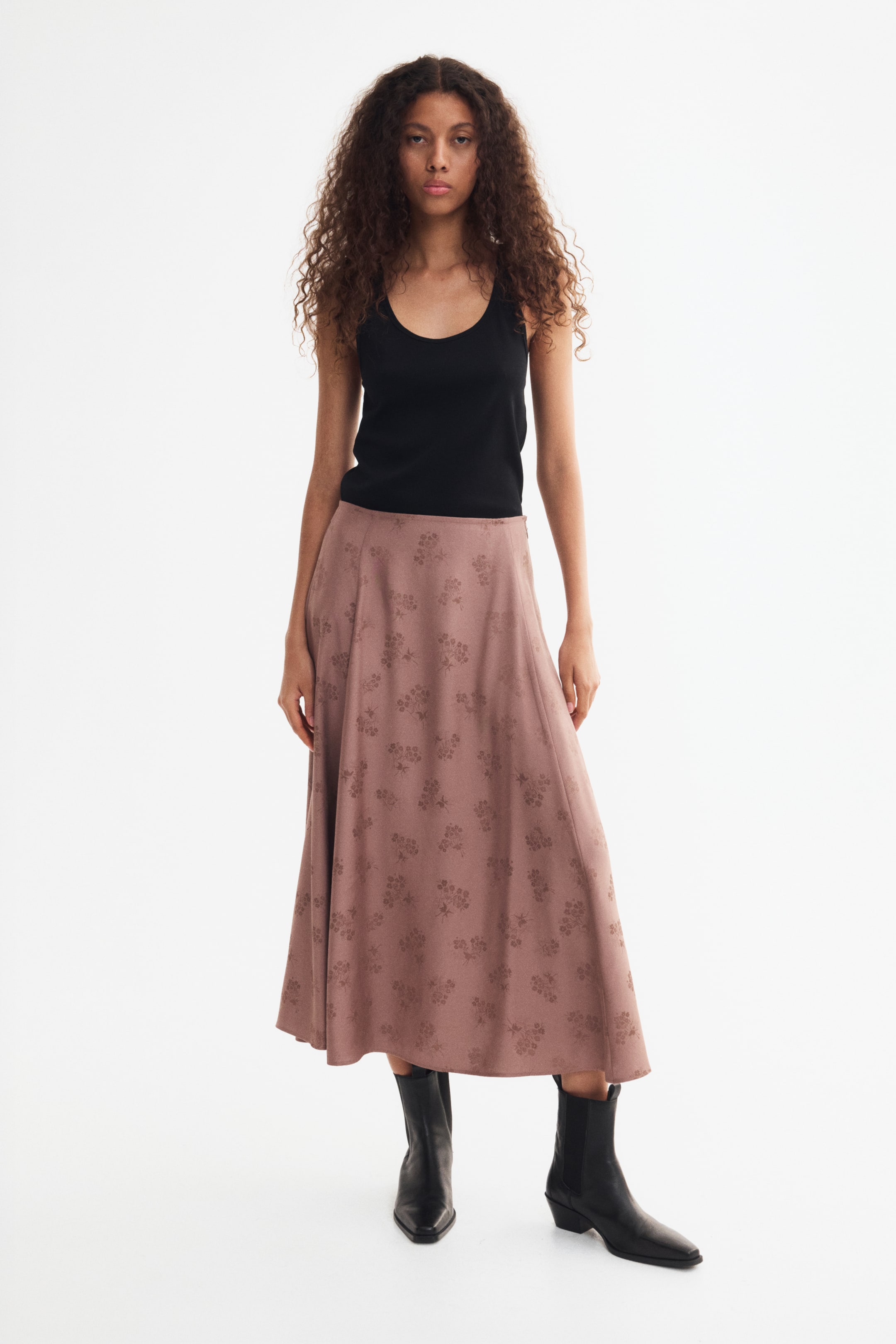 View larger image: A long, A-line skirt in a dusty pink jacquard weave with a subtle floral pattern is being worn by the model, showcasing visible vertical seams down the front. The skirt is being styled with a tucked-in black tank top and black leather boots.