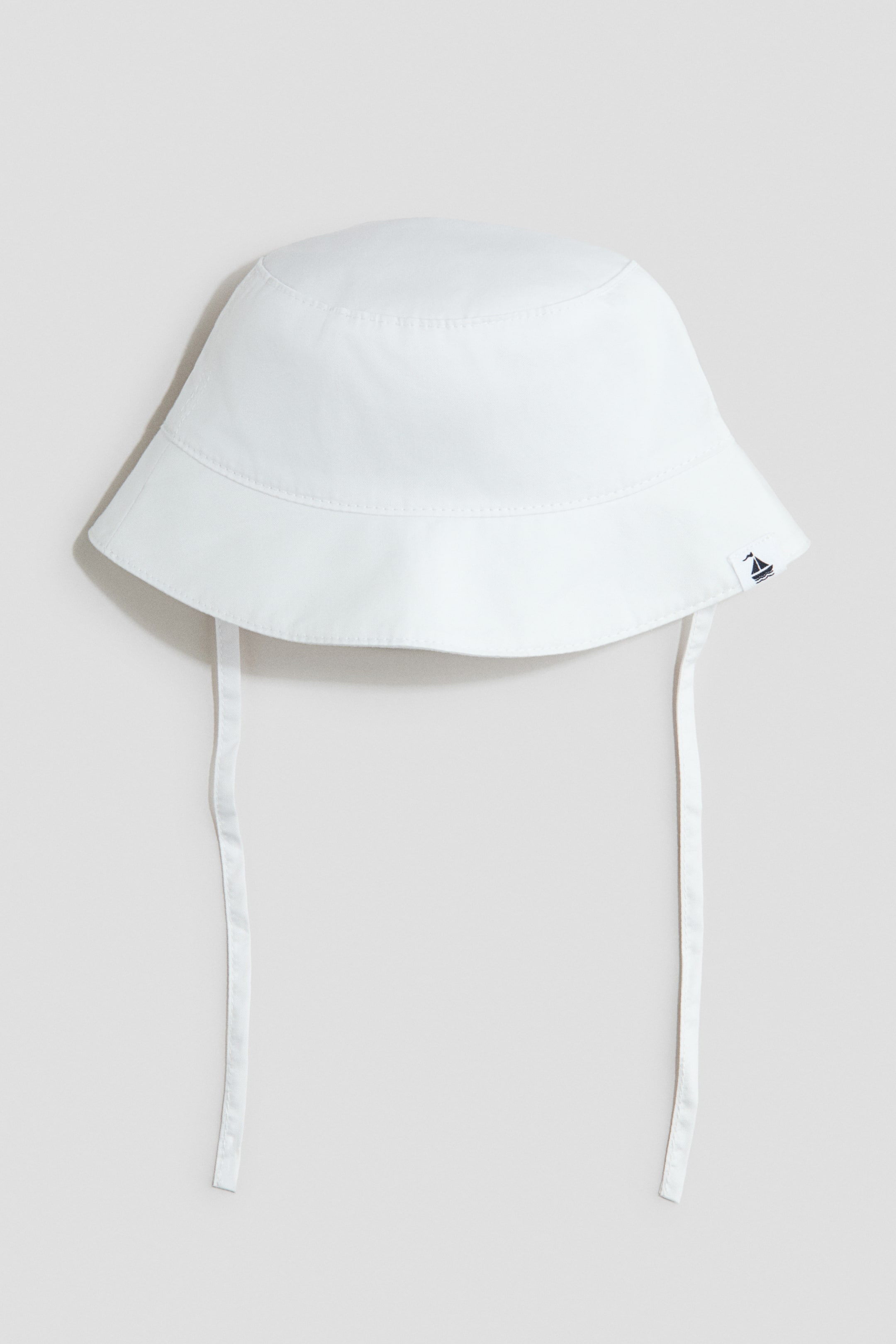 View larger image: A white bucket hat is facing front, crafted from a soft cotton weave. It features ties hanging down from under the chin, and a small sailboat emblem is visible on the right side of the brim.