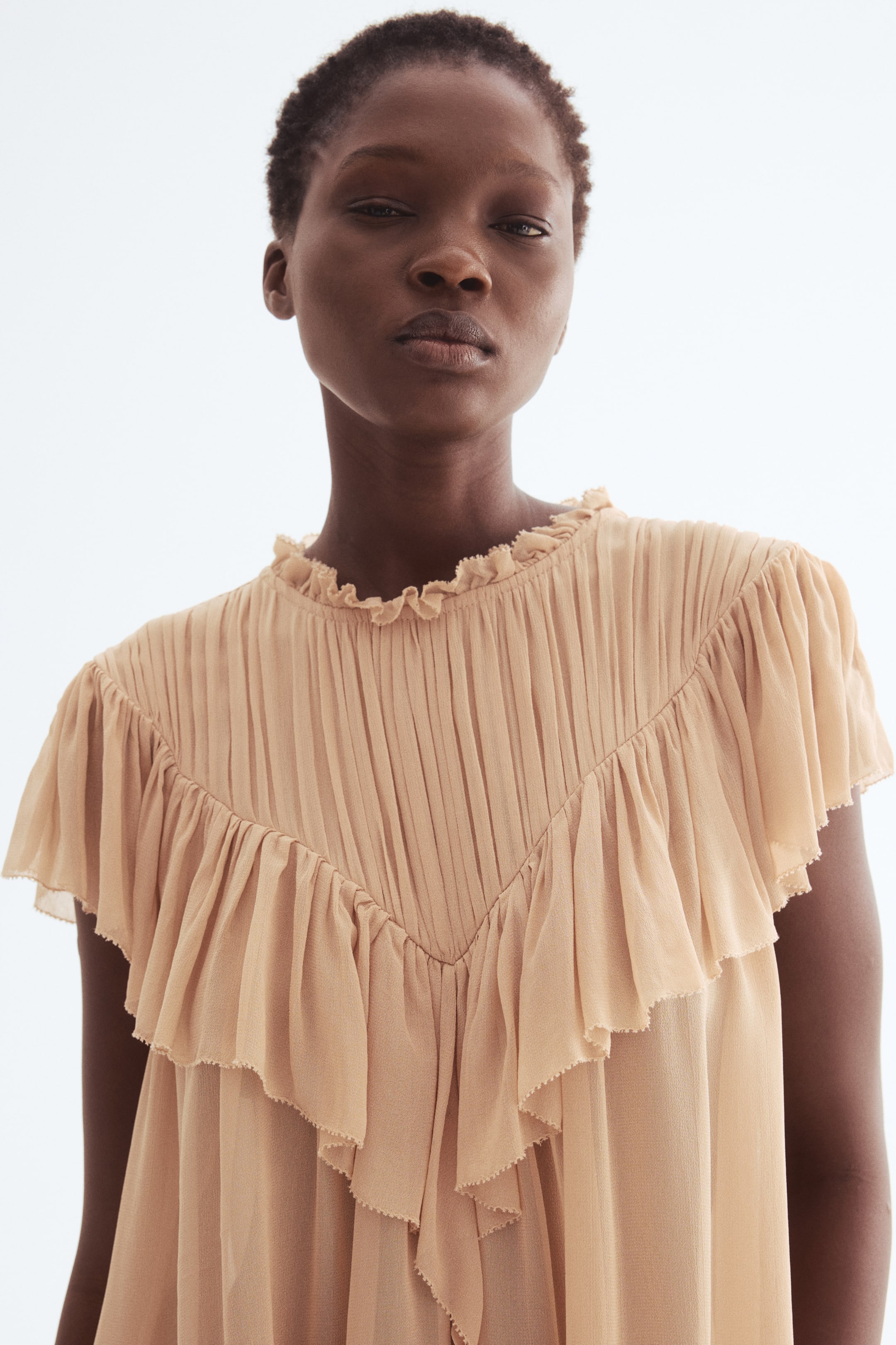 View larger image: A light brown top is featuring a high ruffled neckline, with fine pleats radiating across the chest and shoulders. A V-shaped ruffle overlay is detailing the front, extending into short flutter sleeves.