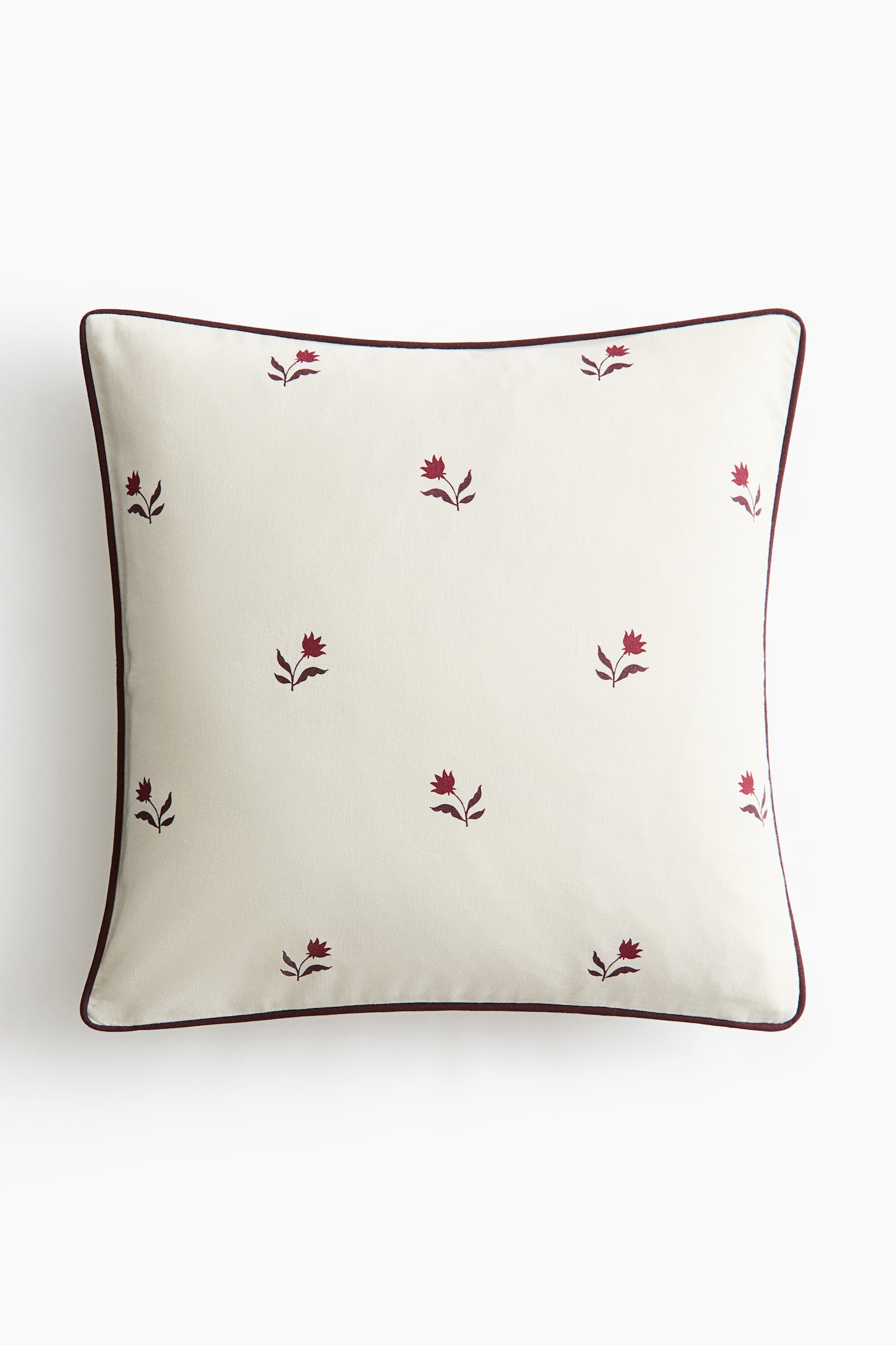 A square throw pillow is facing front, showcasing a cream-coloured fabric adorned with a spaced-out pattern of small, embroidered burgundy floral motifs. The pillow is finished with a thin burgundy piped trim along the outer edges.