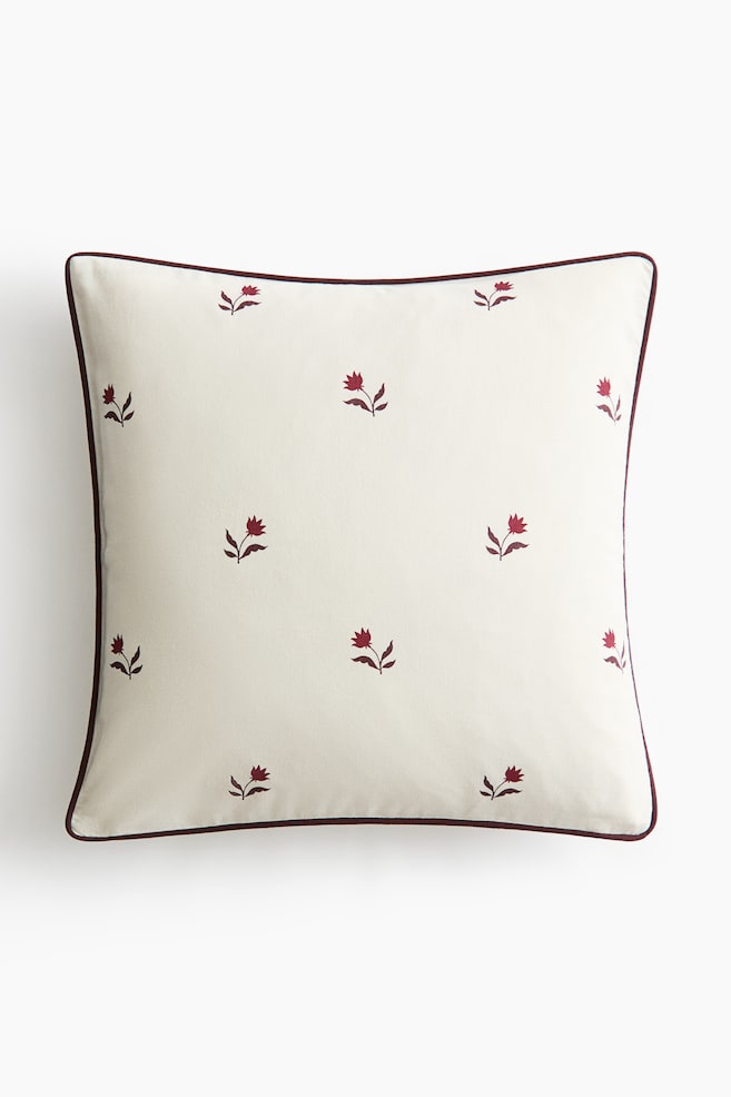 A square throw pillow is shown front-facing, featuring a cream-coloured fabric adorned with a spaced-out pattern of small, embroidered burgundy floral motifs. The pillow is finished with a thin burgundy piped trim along the outer edges. - 1